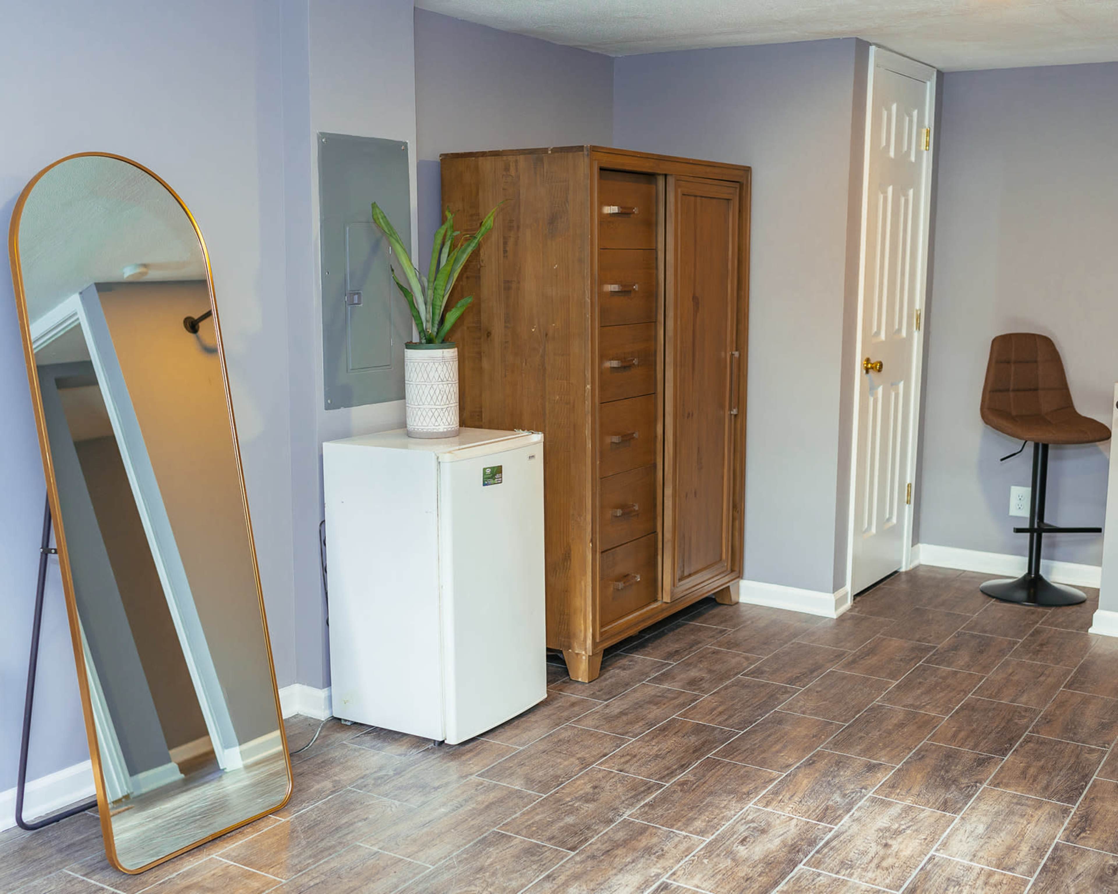 A room with a tall mirror, a wooden dresser, a white refrigerator, and two chairs against a wall with a door.