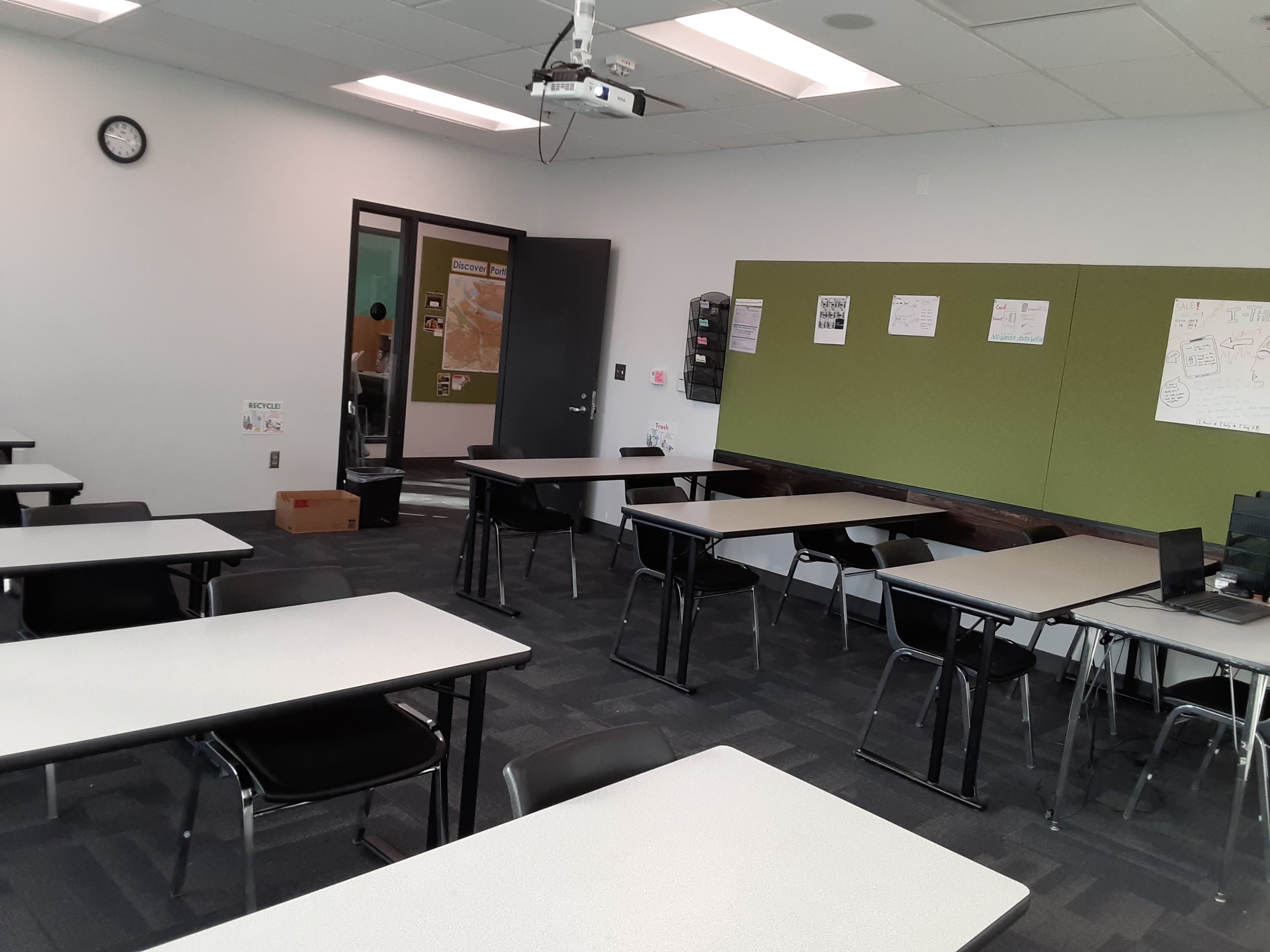 A classroom is set up with several empty tables and chairs, and a projector is mounted on the ceiling.