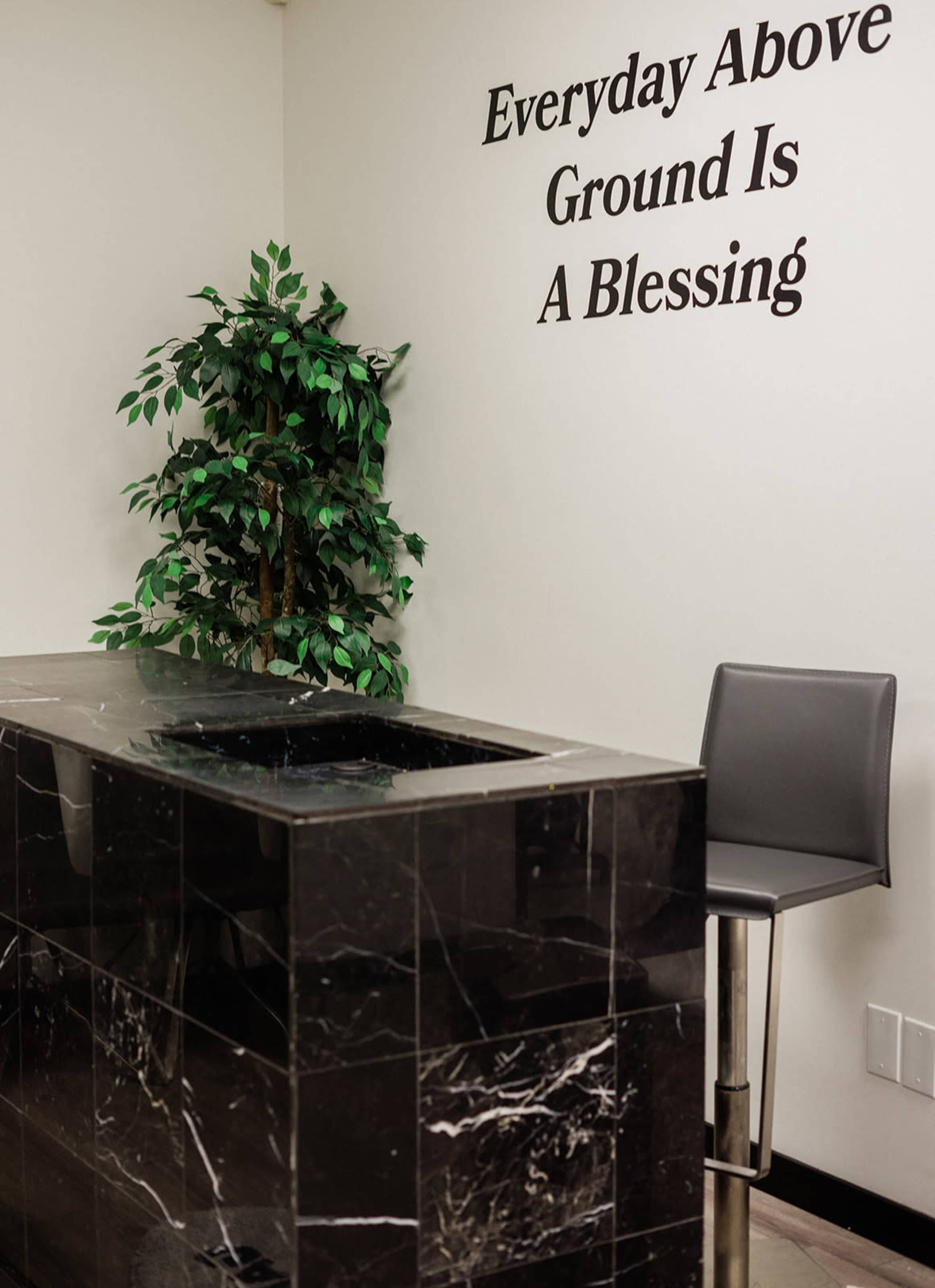 A bar-style seating area is located next to a marble counter, with a faux plant and a wall featuring the phrase "Everyday Above Ground Is A Blessing."