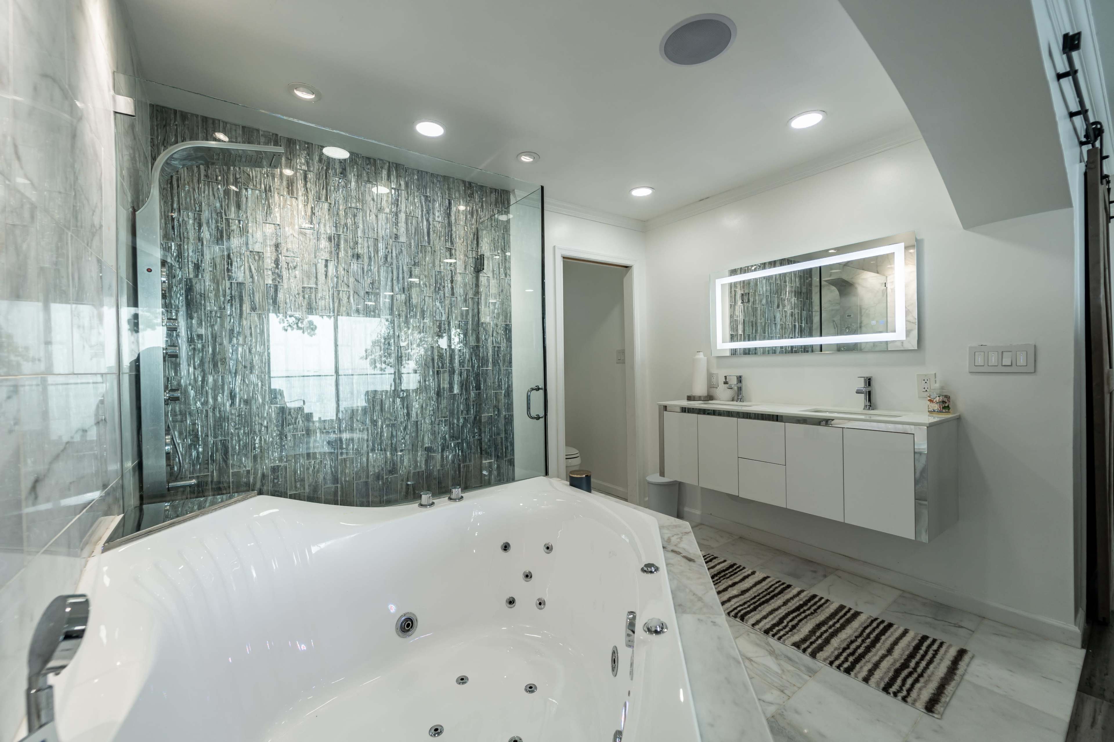 A modern bathroom featuring a glass-enclosed shower, a large jacuzzi tub, a minimalist vanity with a mirror, and marble flooring.
