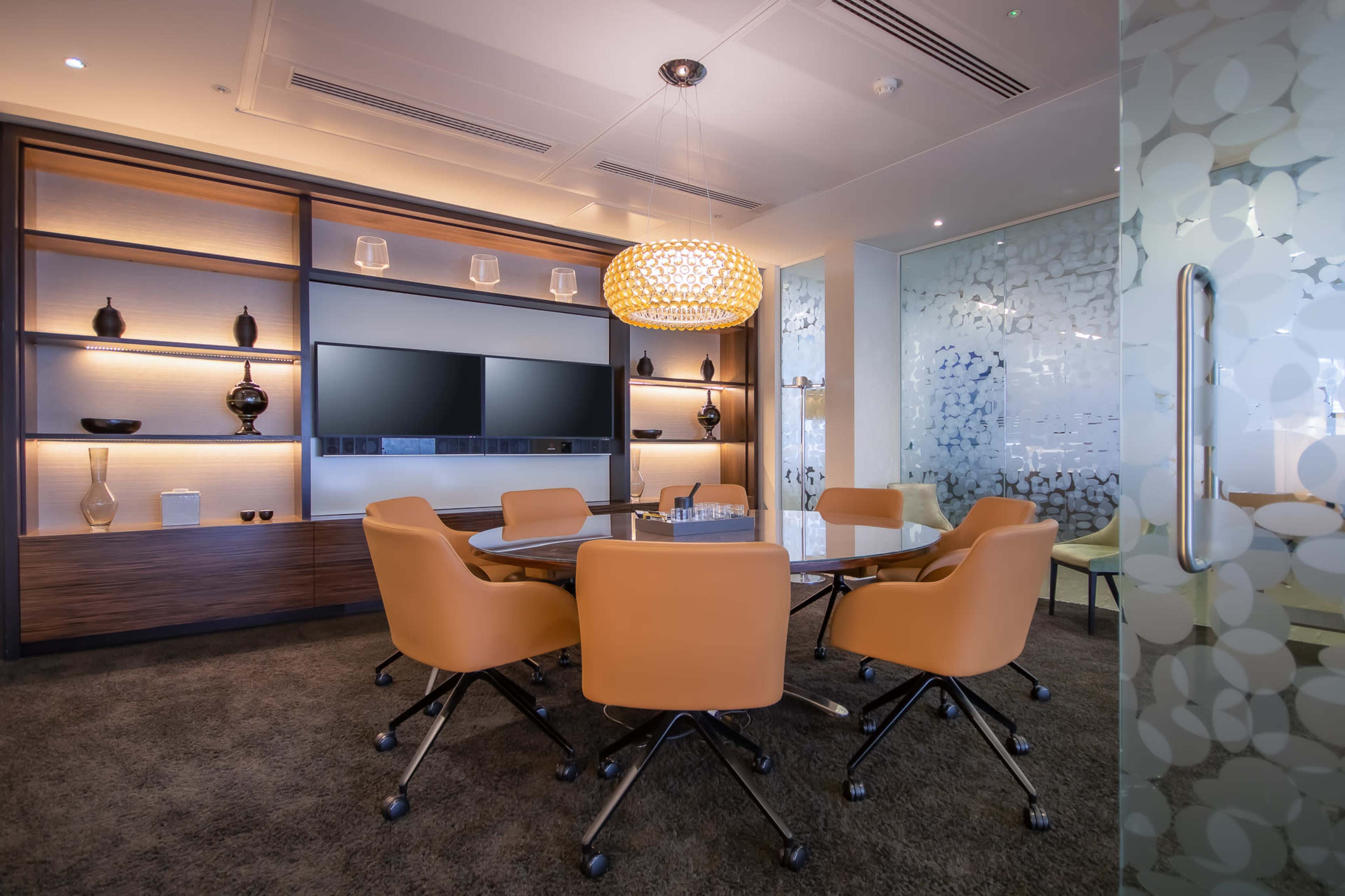 A modern conference room features a round table surrounded by orange chairs, with a display screen and decorative shelving in the background.