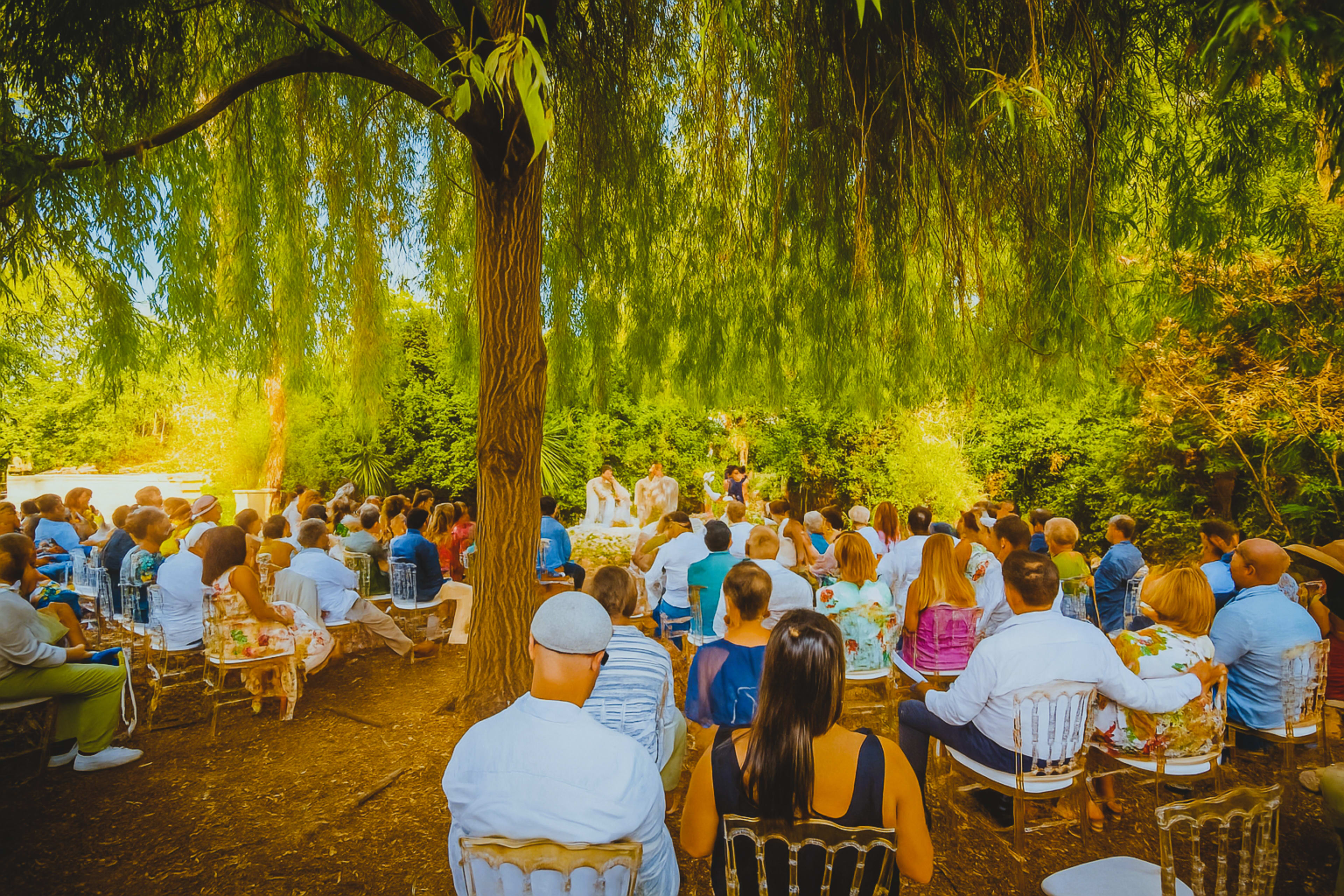 A large group of seated guests gather under a willow tree for an outdoor wedding ceremony.
