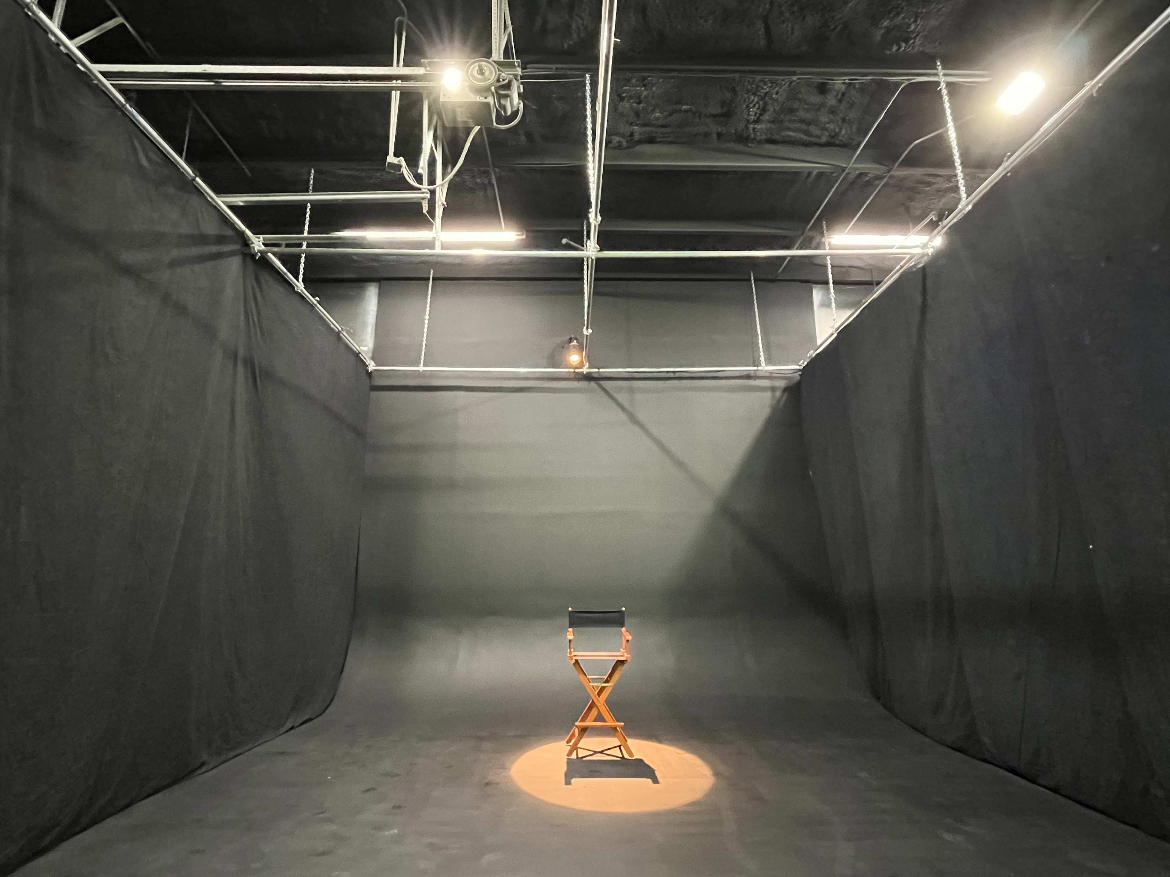 A single wooden director's chair is illuminated on the floor of a darkened studio surrounded by black curtains.