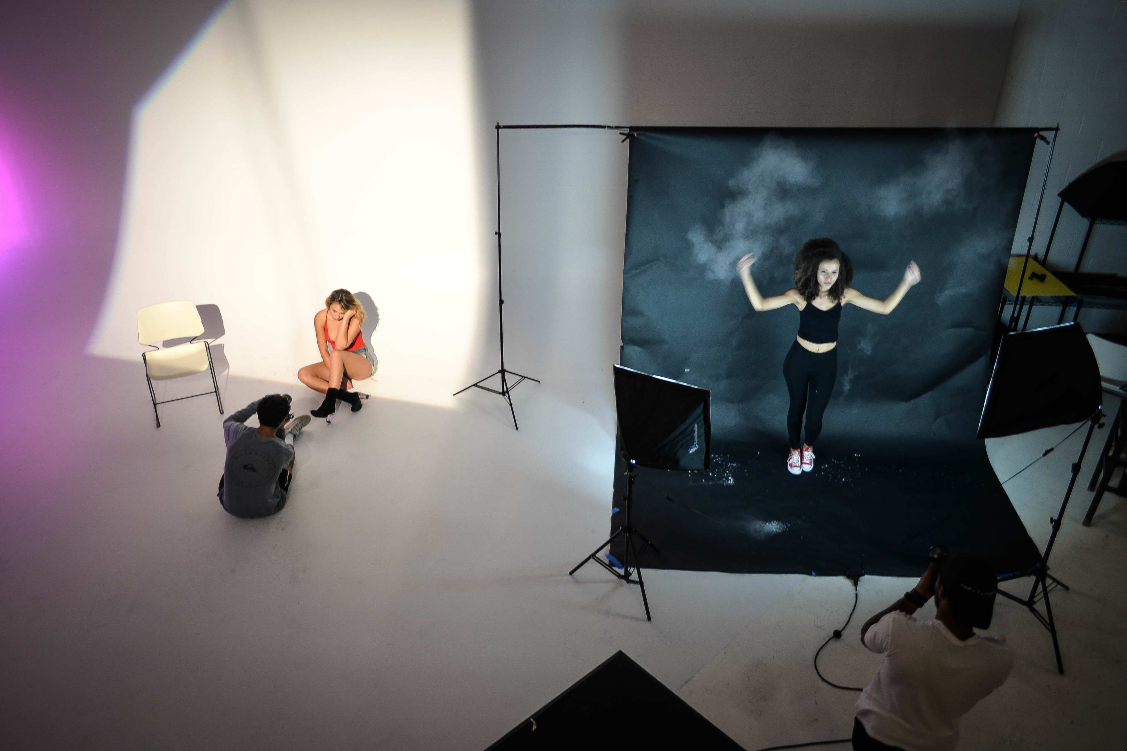 In a photography studio, one model poses in front of a black backdrop while another sits on a chair, and a photographer takes shots from a low angle.