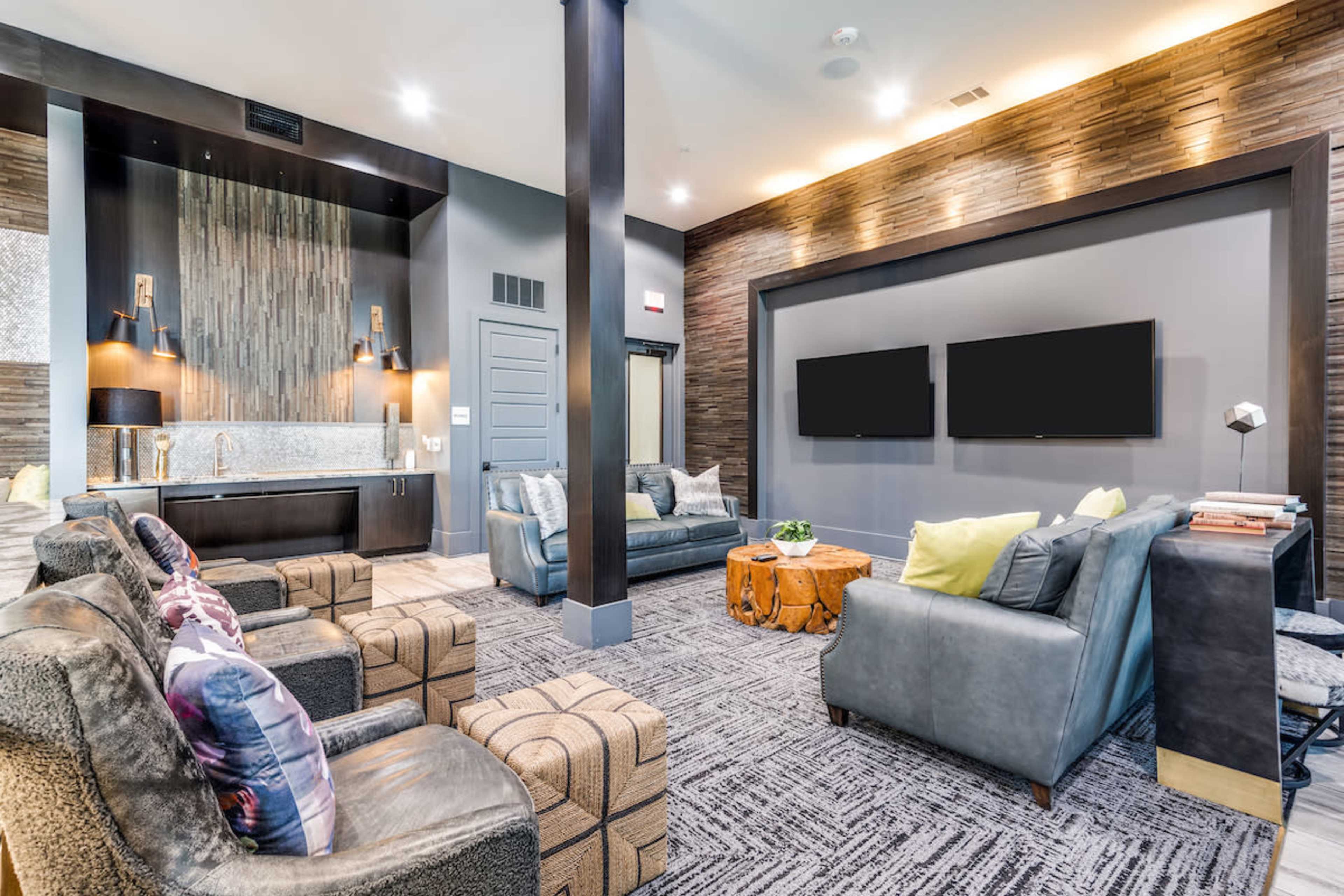 A modern lounge area with two sofas, armchairs, and two wall-mounted TVs, featuring a wooden accent wall and a kitchenette in the background.