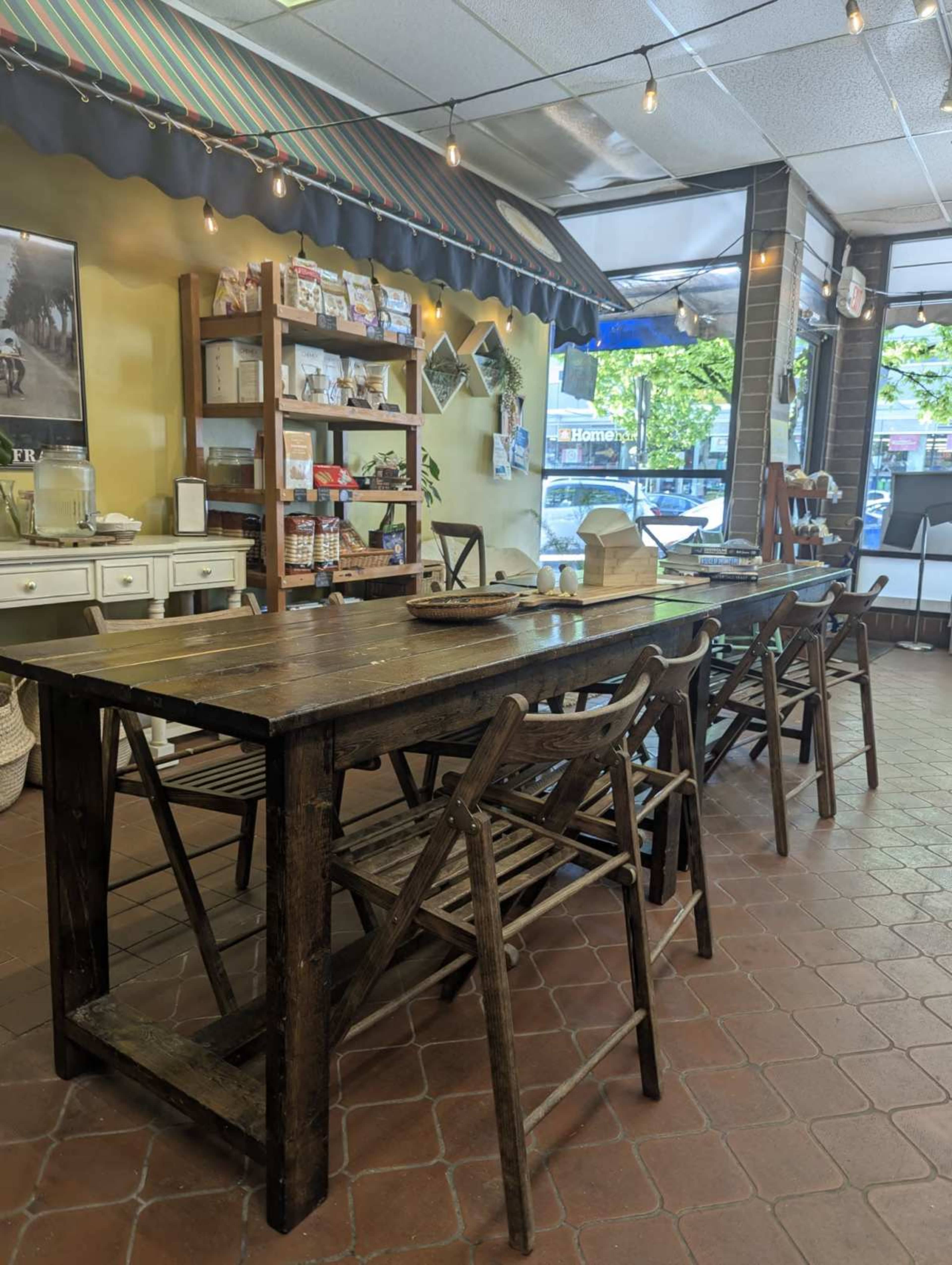The image shows a rustic wooden table surrounded by folding chairs in a shop with shelves displaying various items in the background.
