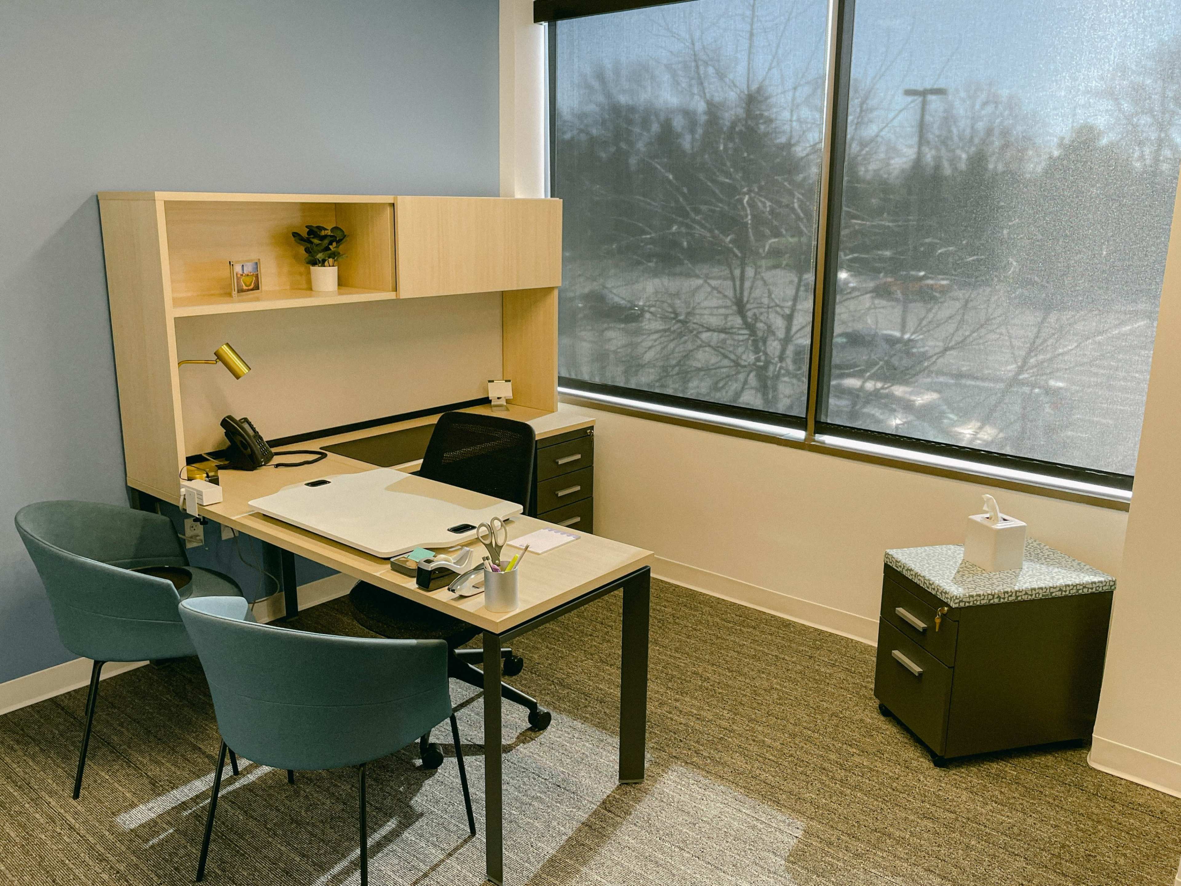 A minimalist office space features a desk with a chair, a small plant, a phone, and a filing cabinet near a large window letting in natural light.