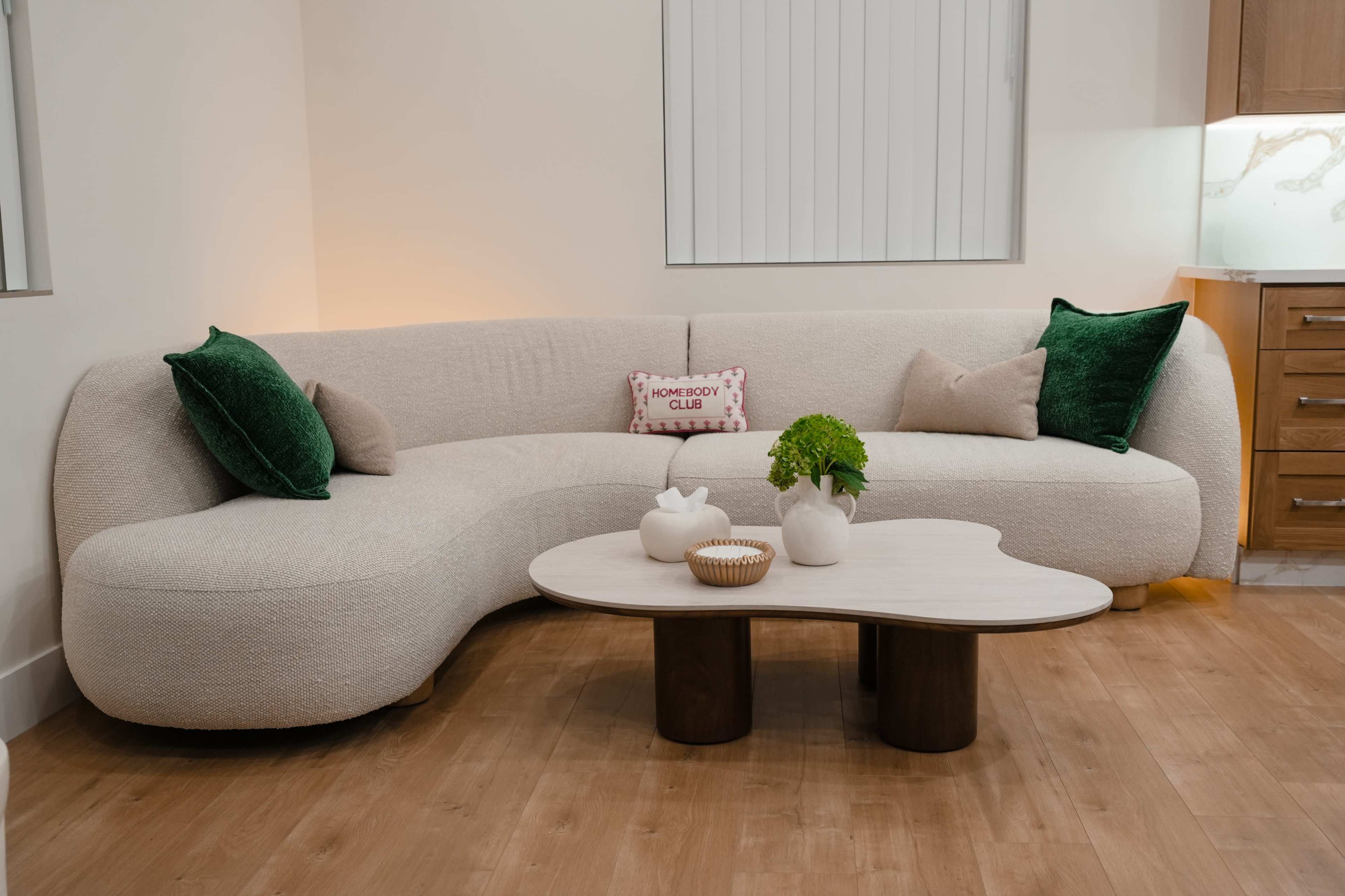 A curved beige sofa with green cushions sits beside a round wooden coffee table in a well-lit living room.
