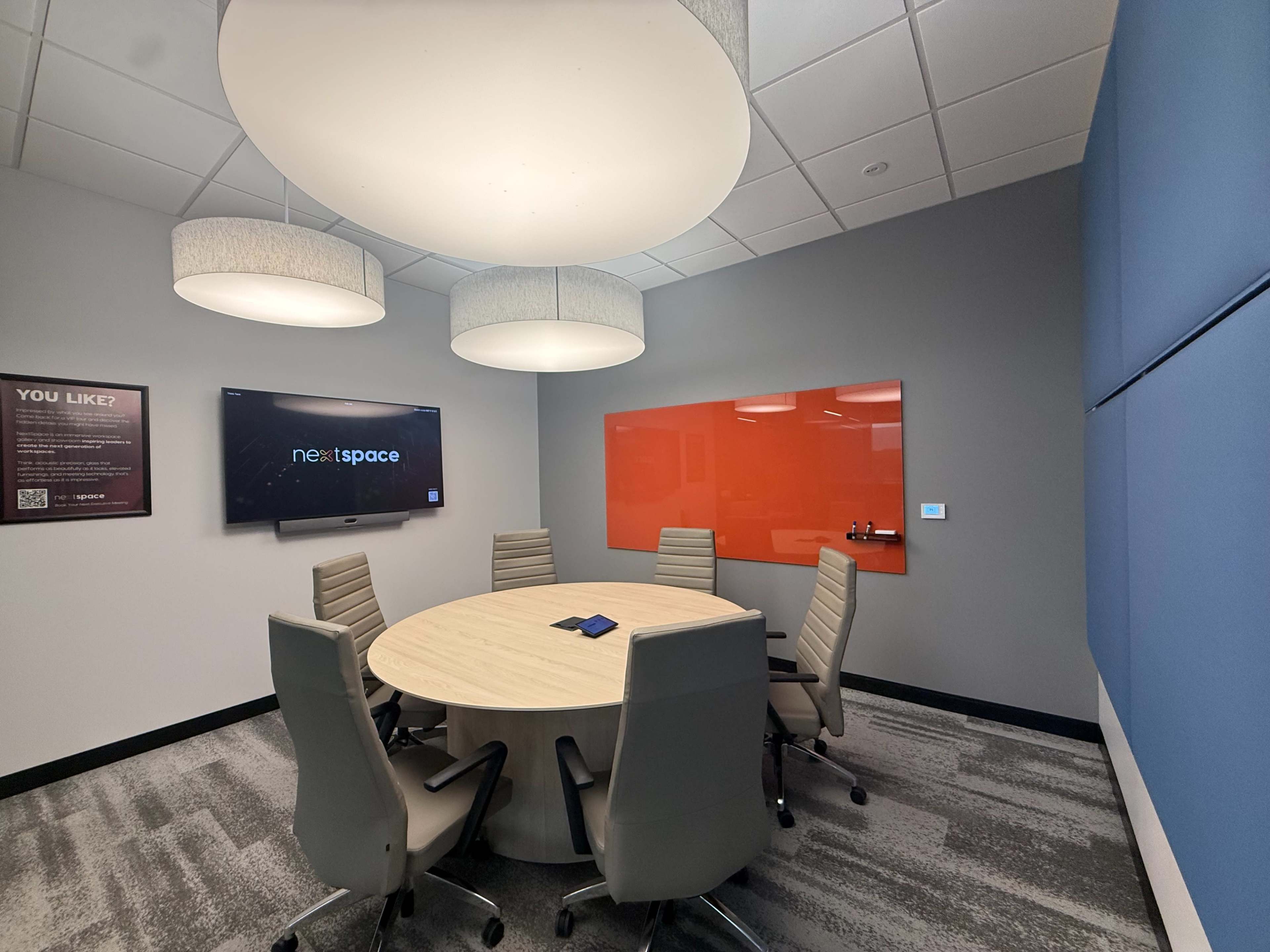 The image shows a modern conference room equipped with a round table, six ergonomic chairs, and a large screen mounted on the wall, featuring a bright orange accent panel.