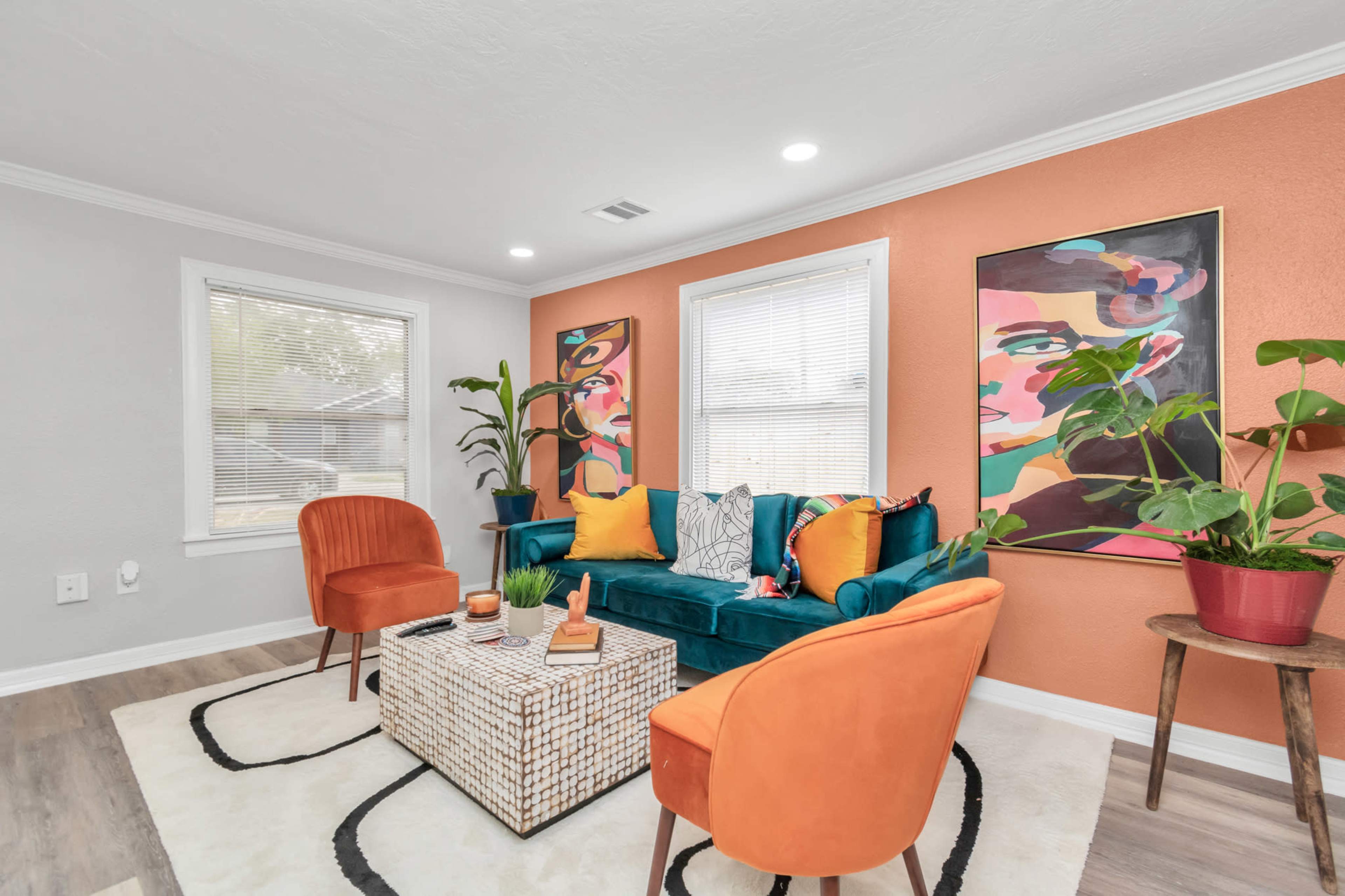 A living room with a teal sofa, two orange chairs, a patterned coffee table, and colorful artwork on the walls.