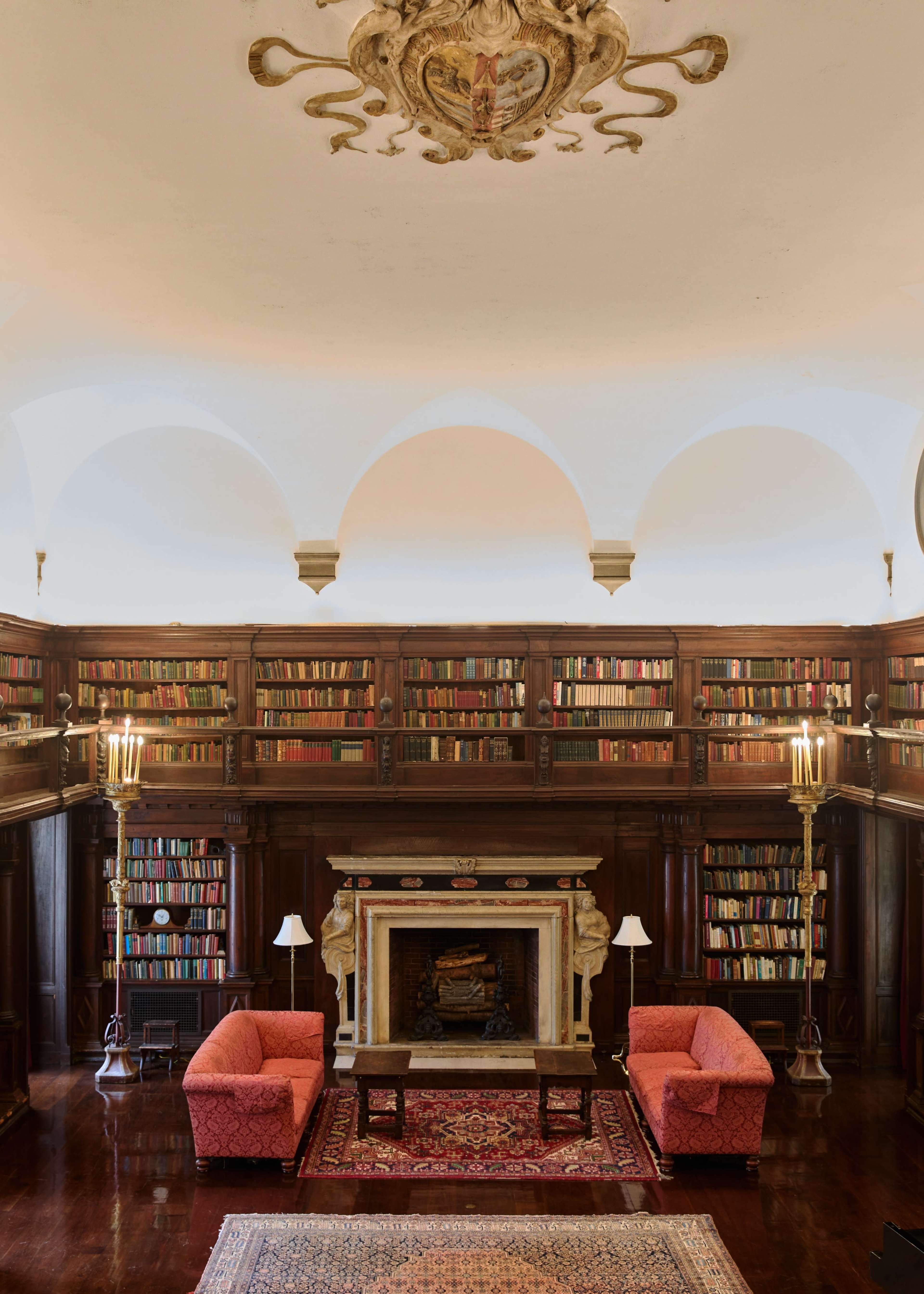 The image shows an elegant library with tall bookshelves filled with books, a central fireplace, and two upholstered chairs arranged on a patterned rug.