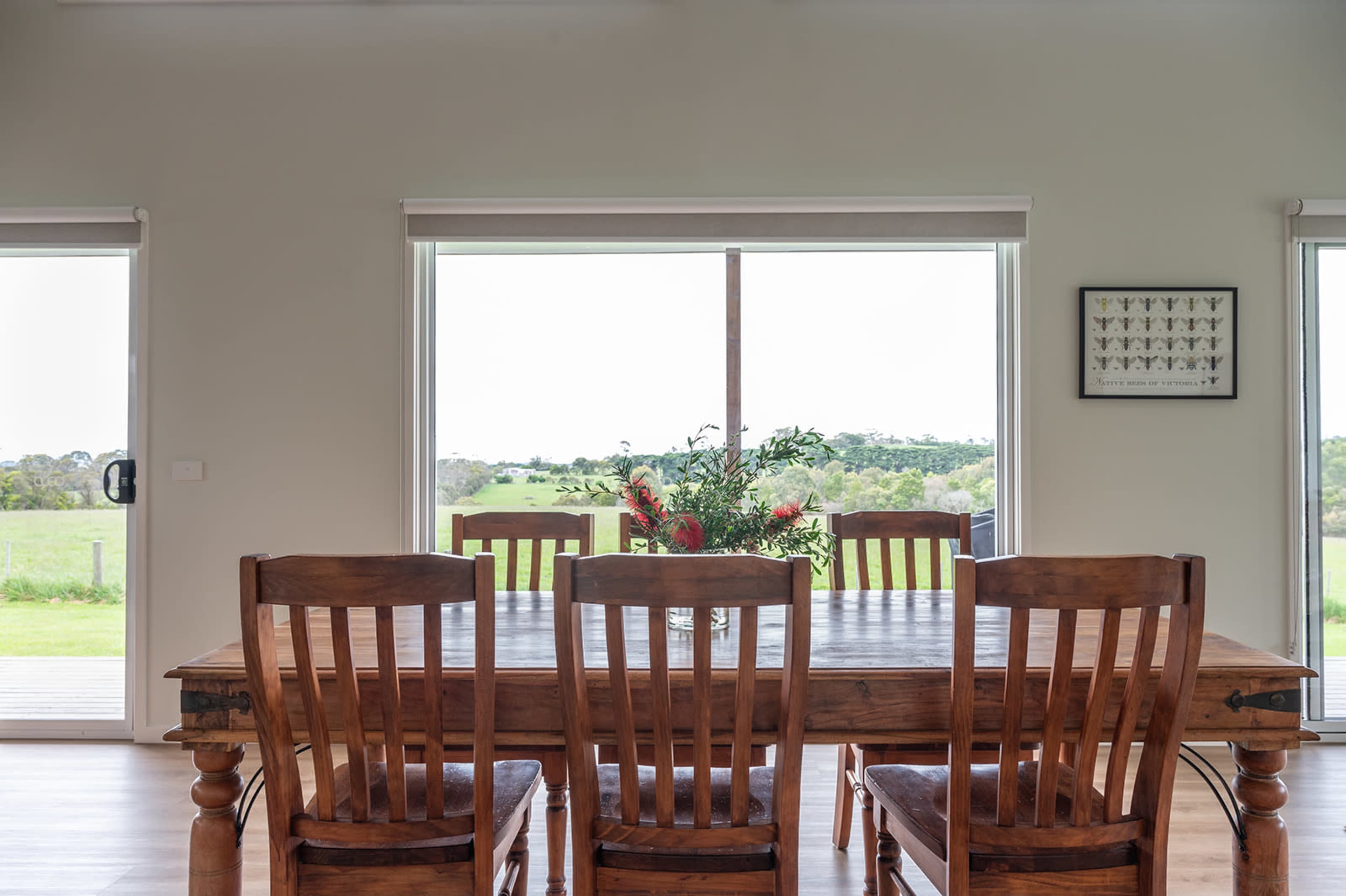 A wooden dining table with six chairs is set in a room with large windows overlooking a green landscape.