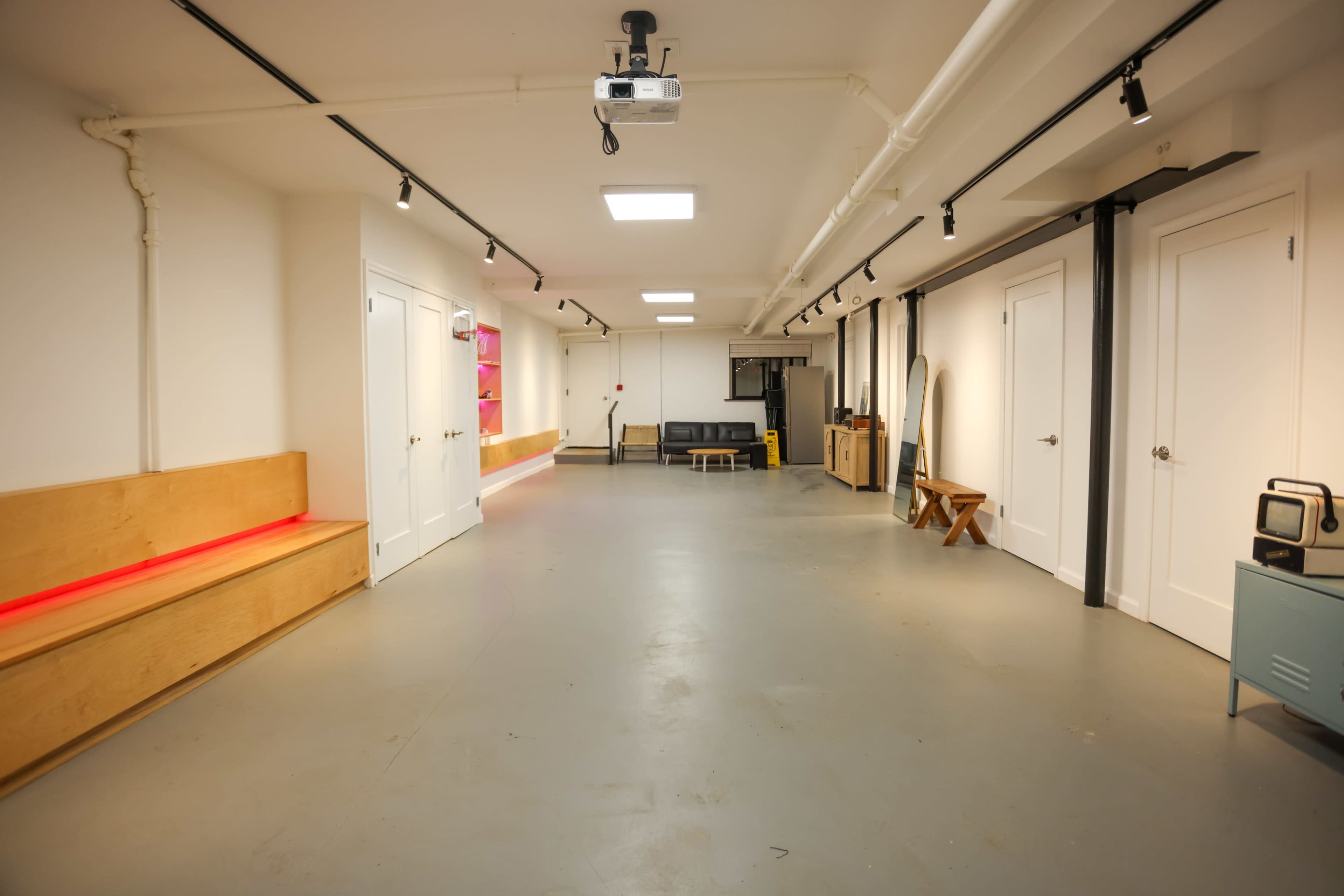 The image shows a spacious, minimalistic room with concrete flooring, white walls, and various furniture against one side, including a sofa and a small table.