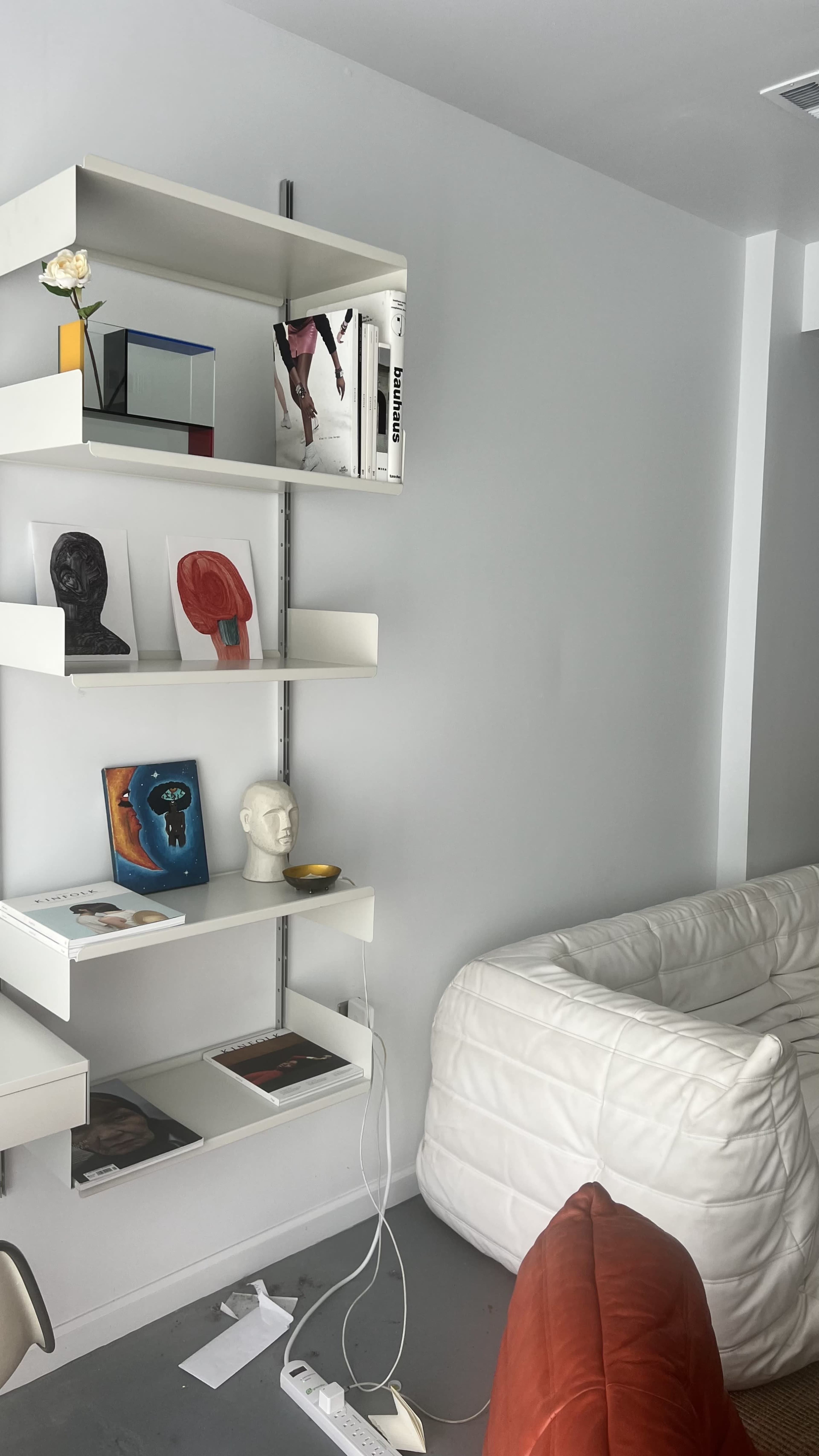 The image shows a modern white shelf with various art books and decorative items, alongside a white couch with orange cushions and a power strip on the floor.