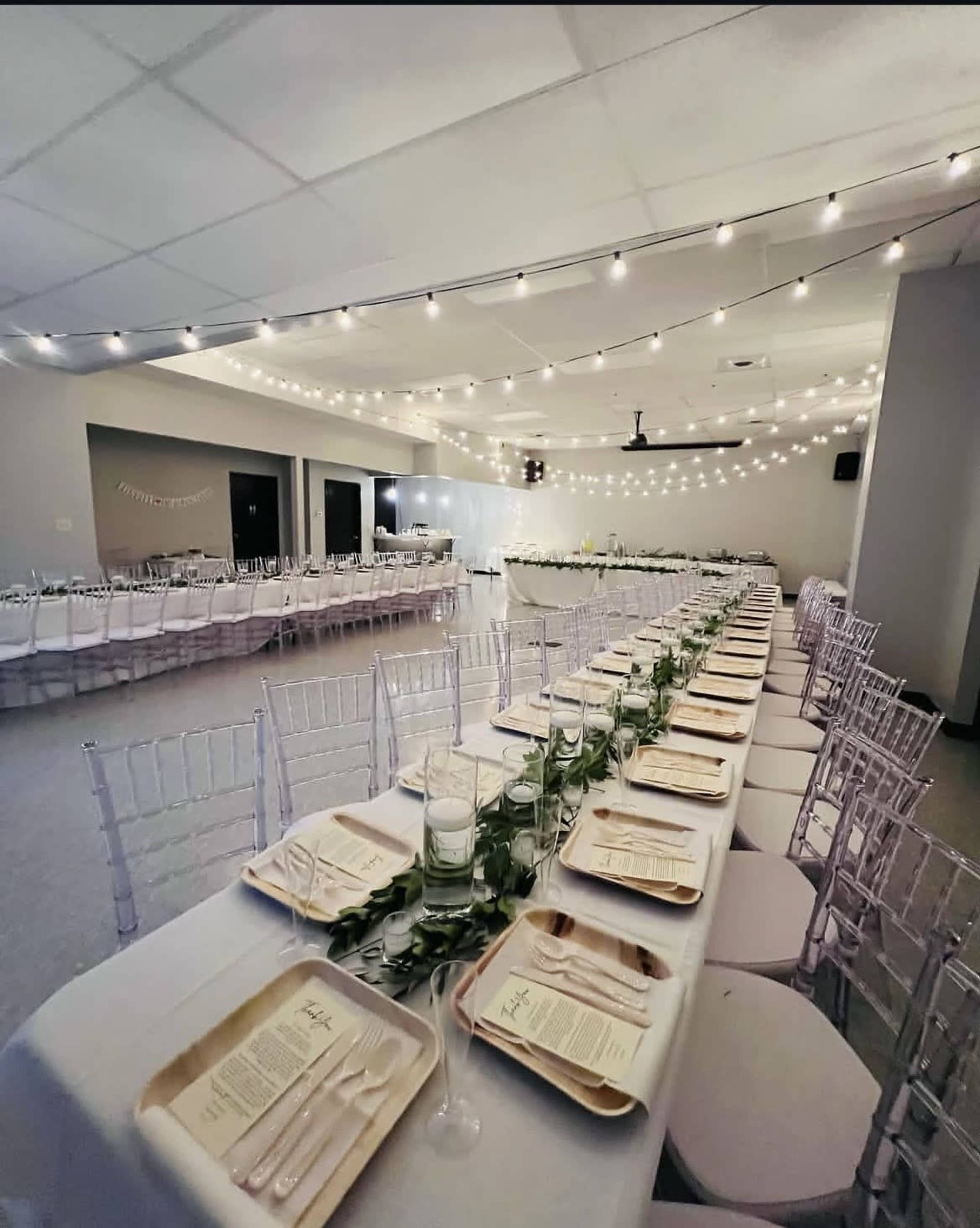 A large banquet hall is set for an elegant dinner, featuring multiple long tables with white tablecloths, chiavari chairs, and string lights overhead.
