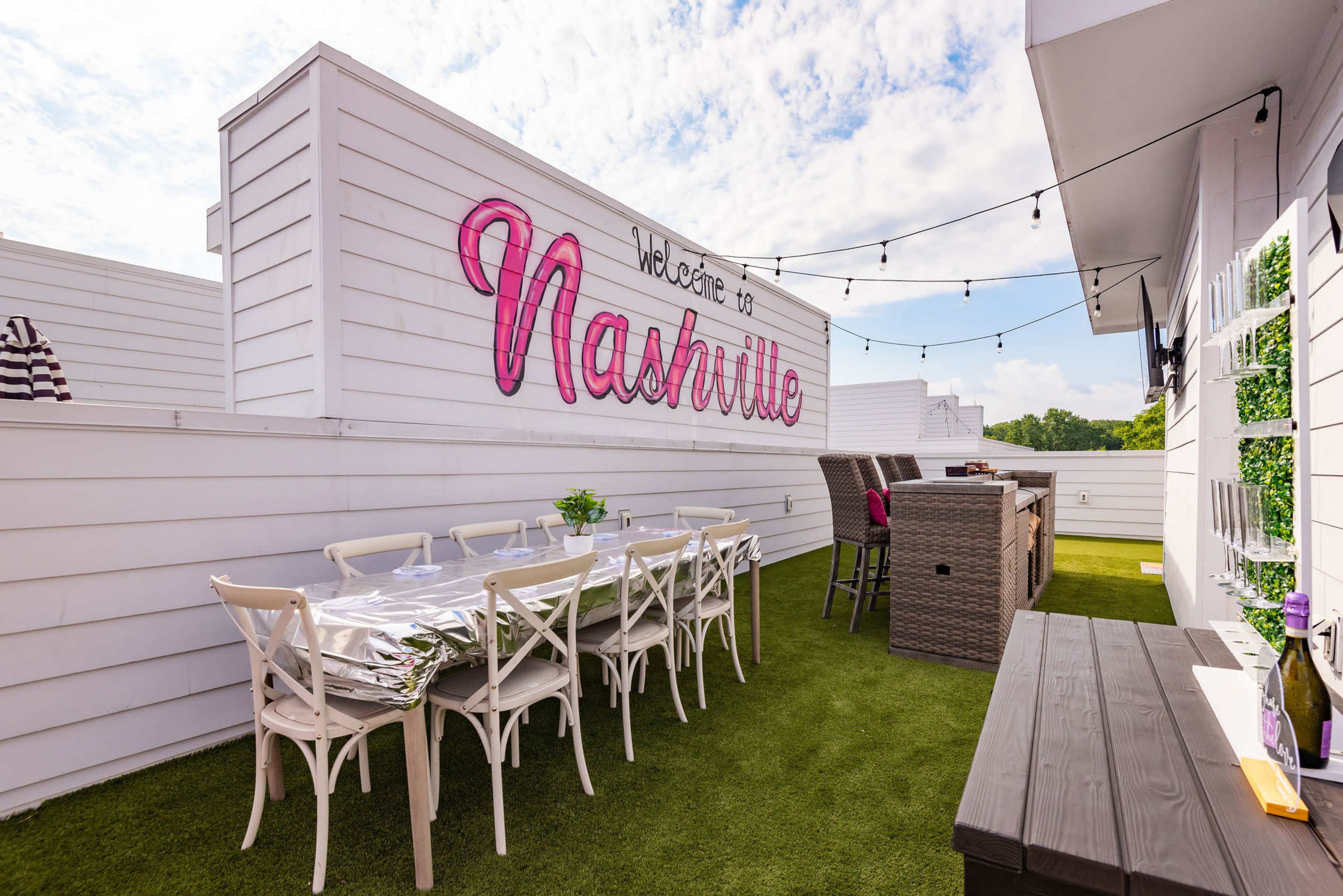 The image shows a rooftop terrace in Nashville featuring white walls, string lights, a long dining table, and wicker furniture.