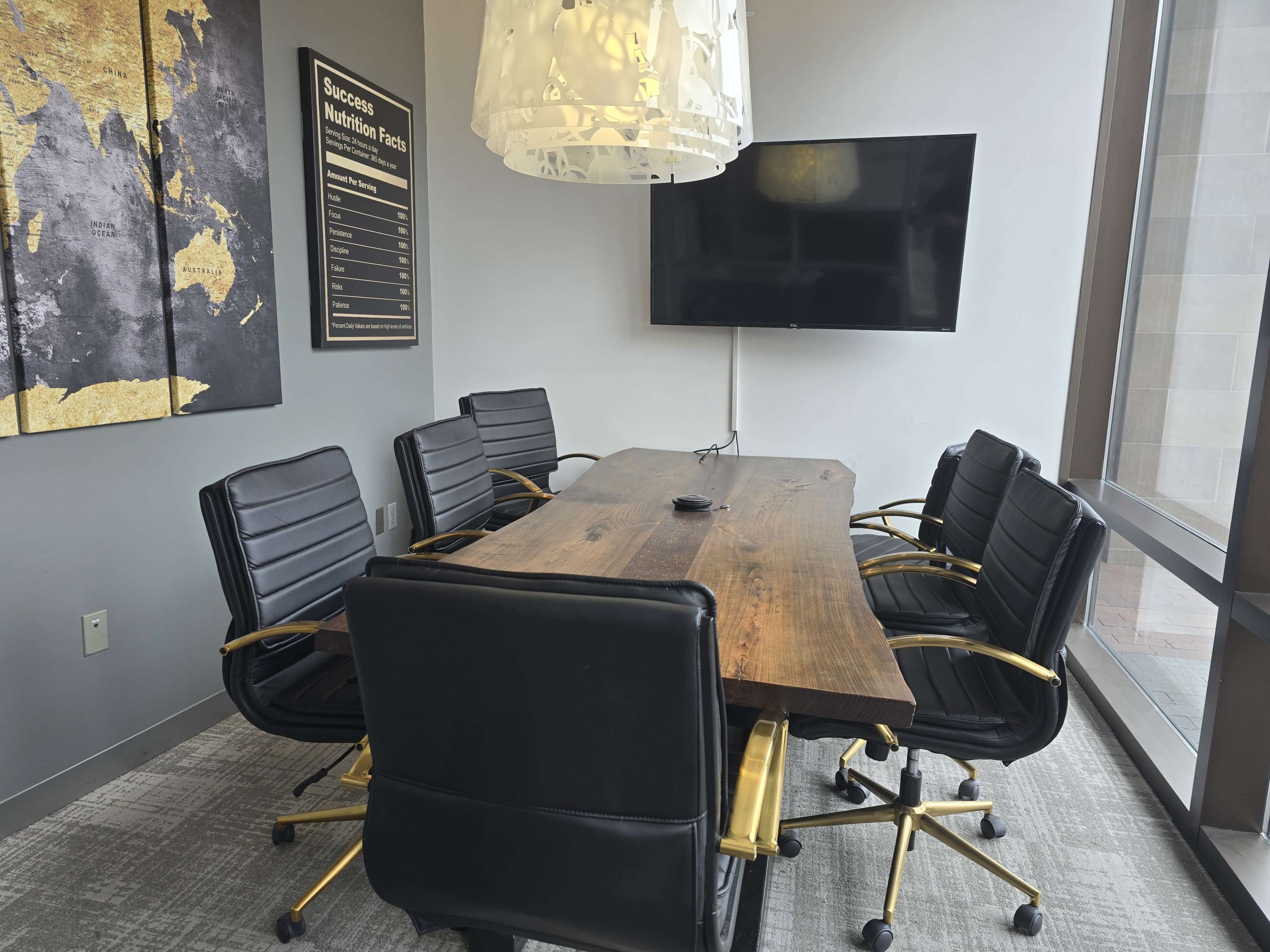 A conference room features a wooden table surrounded by six black leather chairs, with a large television mounted on the wall and decorative artwork in the background.