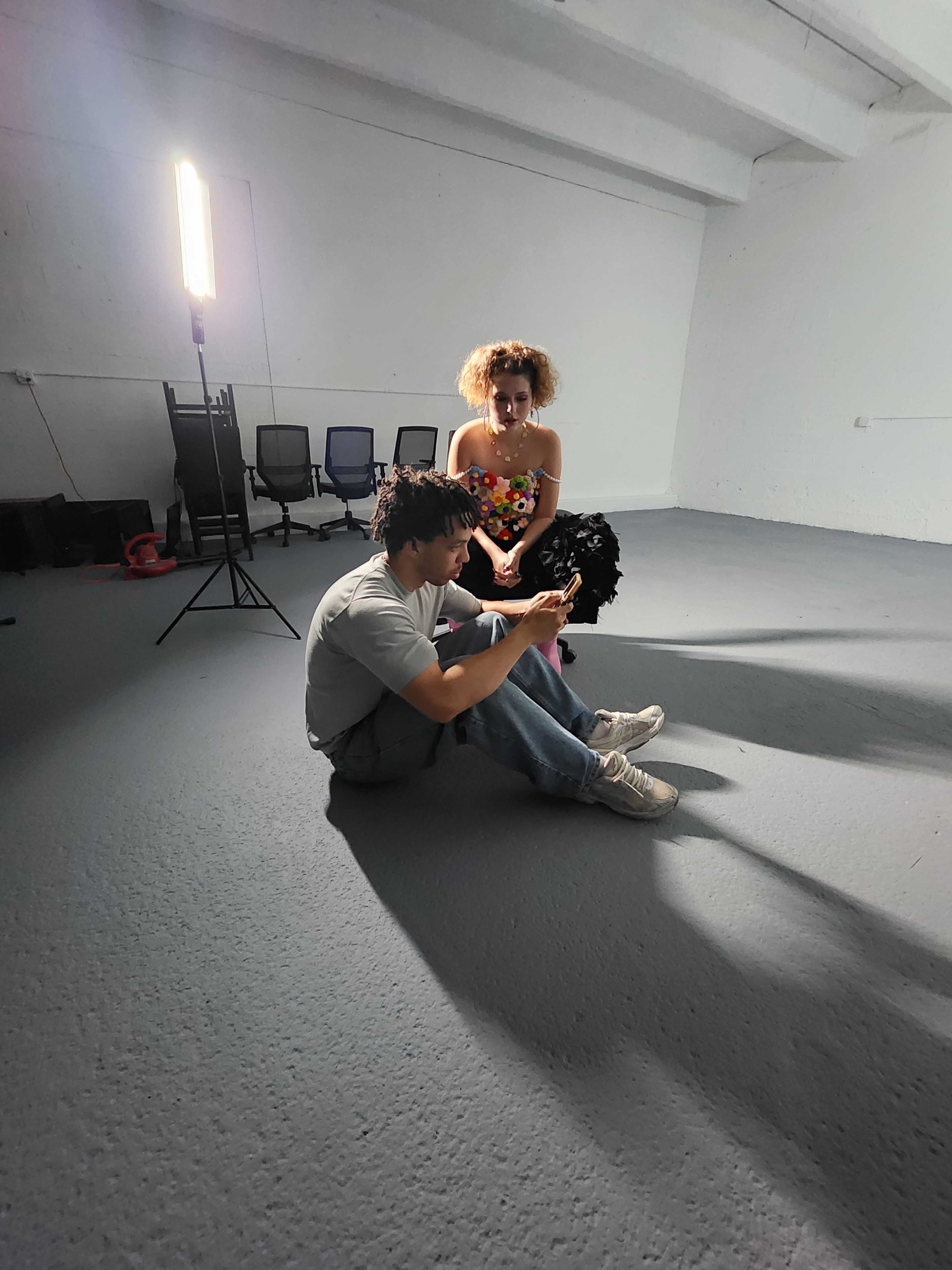A woman in a colorful dress sits on the floor while a man beside her looks at his phone in a well-lit, empty room.