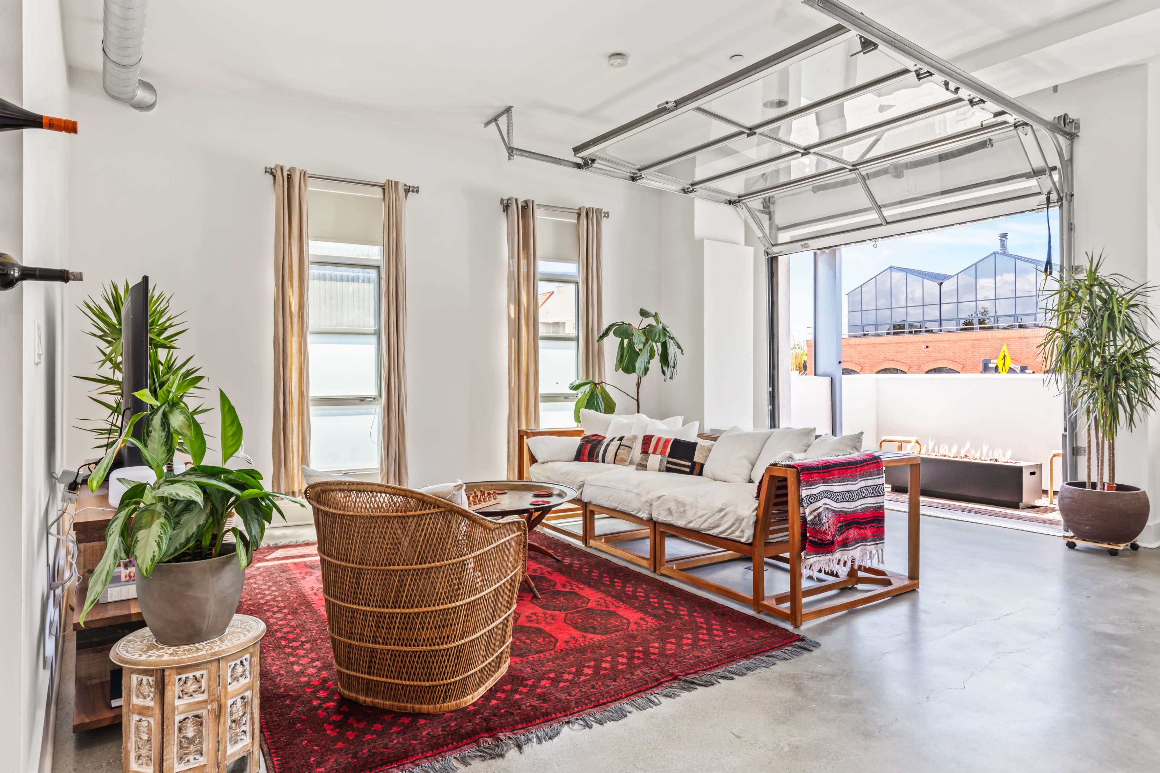 The image shows a modern living space featuring a comfortable seating area with a patterned rug, large windows, and indoor plants.