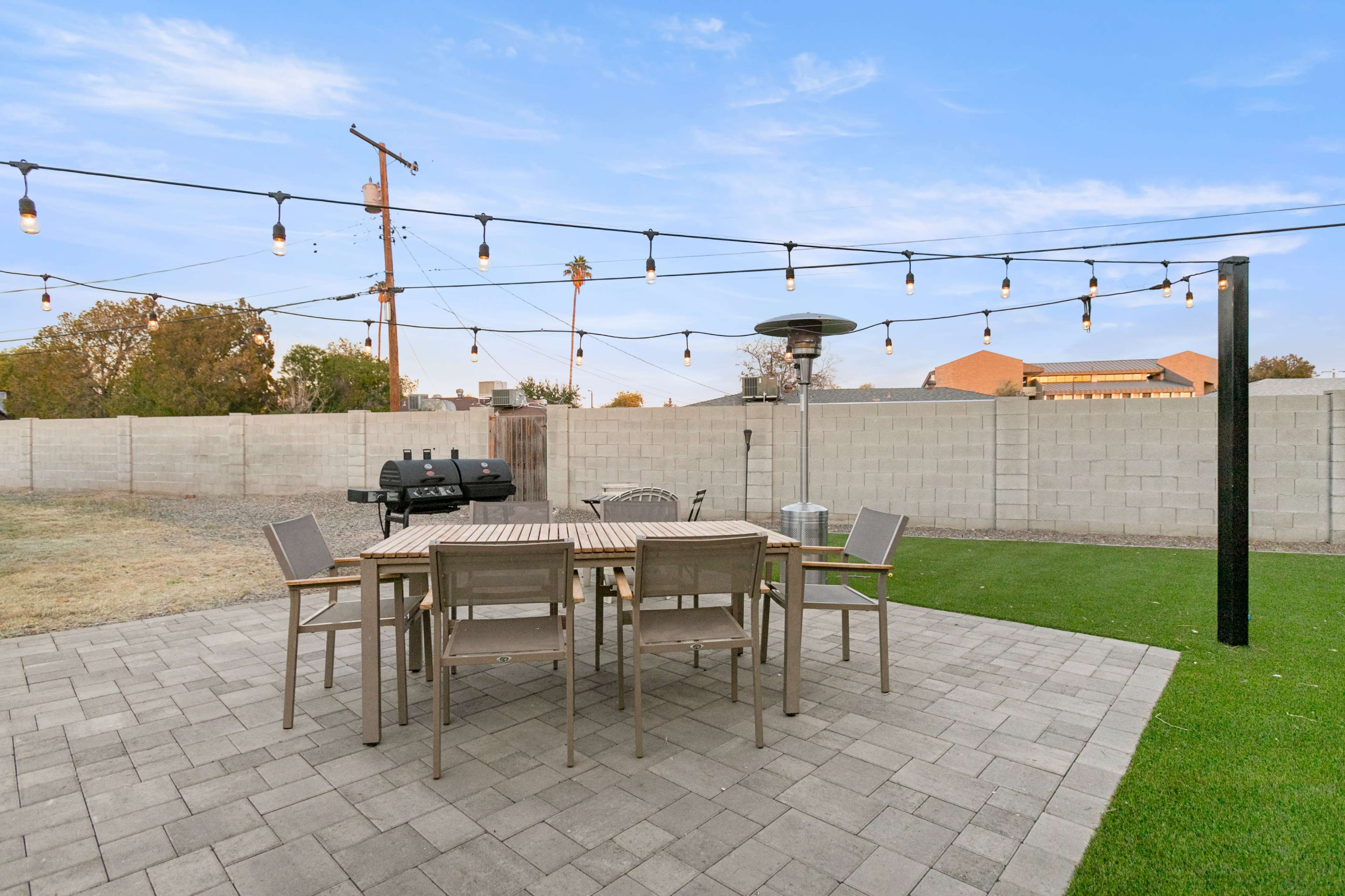 A patio area features a wooden dining table surrounded by chairs, with string lights overhead and a gas grill in the background.
