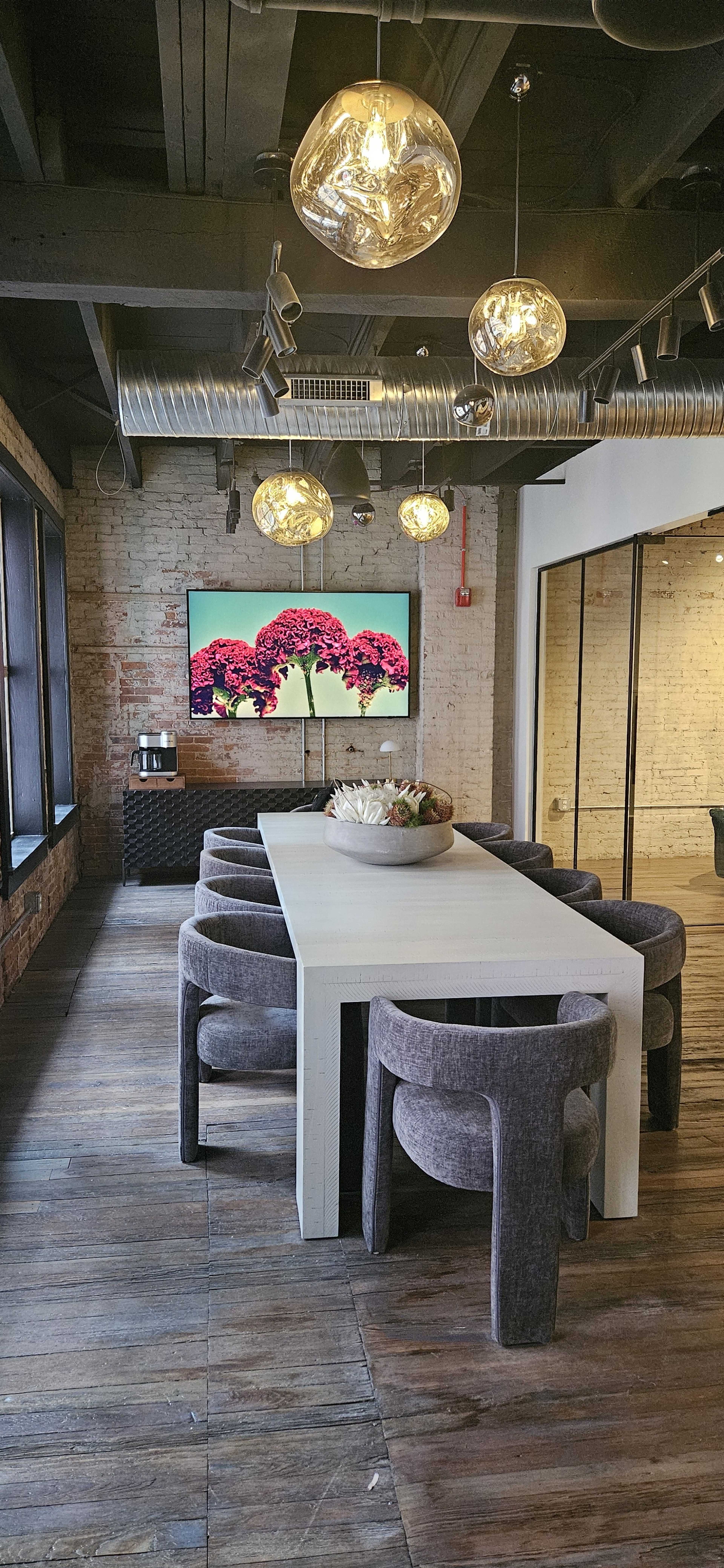 A long, white table surrounded by gray chairs is lit by hanging pendant lights in a modern conference room featuring a large screen displaying a floral image.