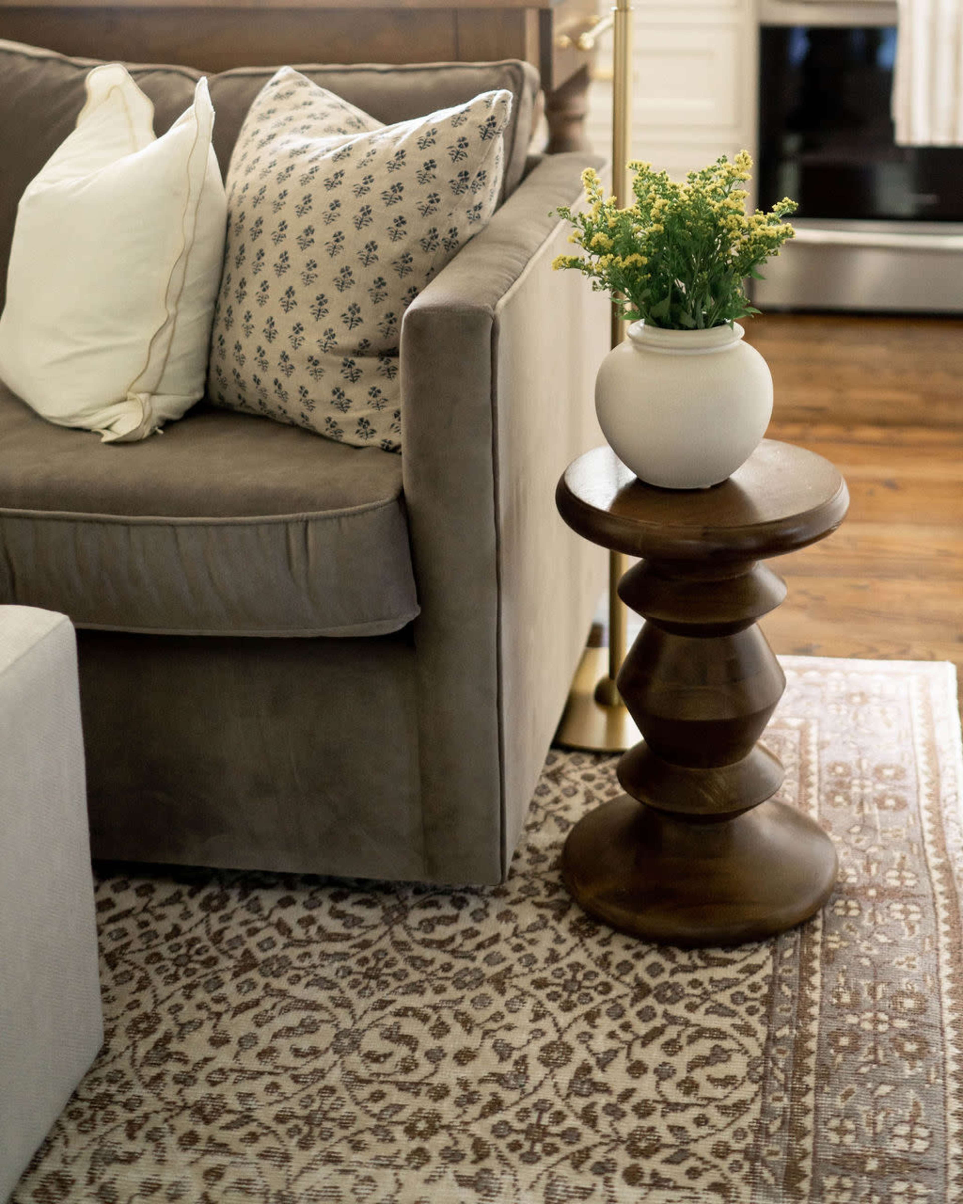 A gray sofa with patterned pillows sits next to a wooden side table holding a potted plant, all placed on a decorative rug.