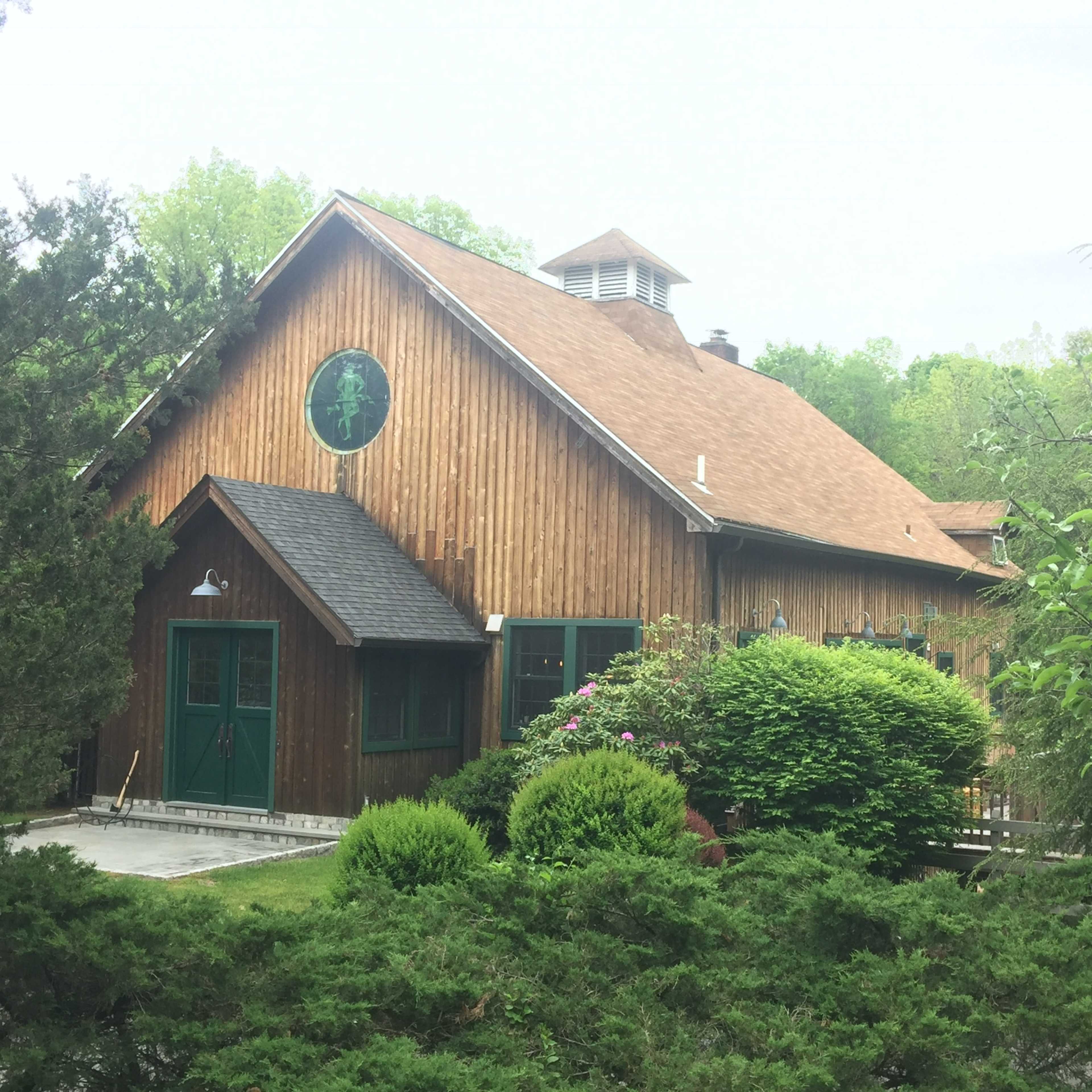 A wooden barn-style building with green doors and windows is surrounded by shrubs and trees.