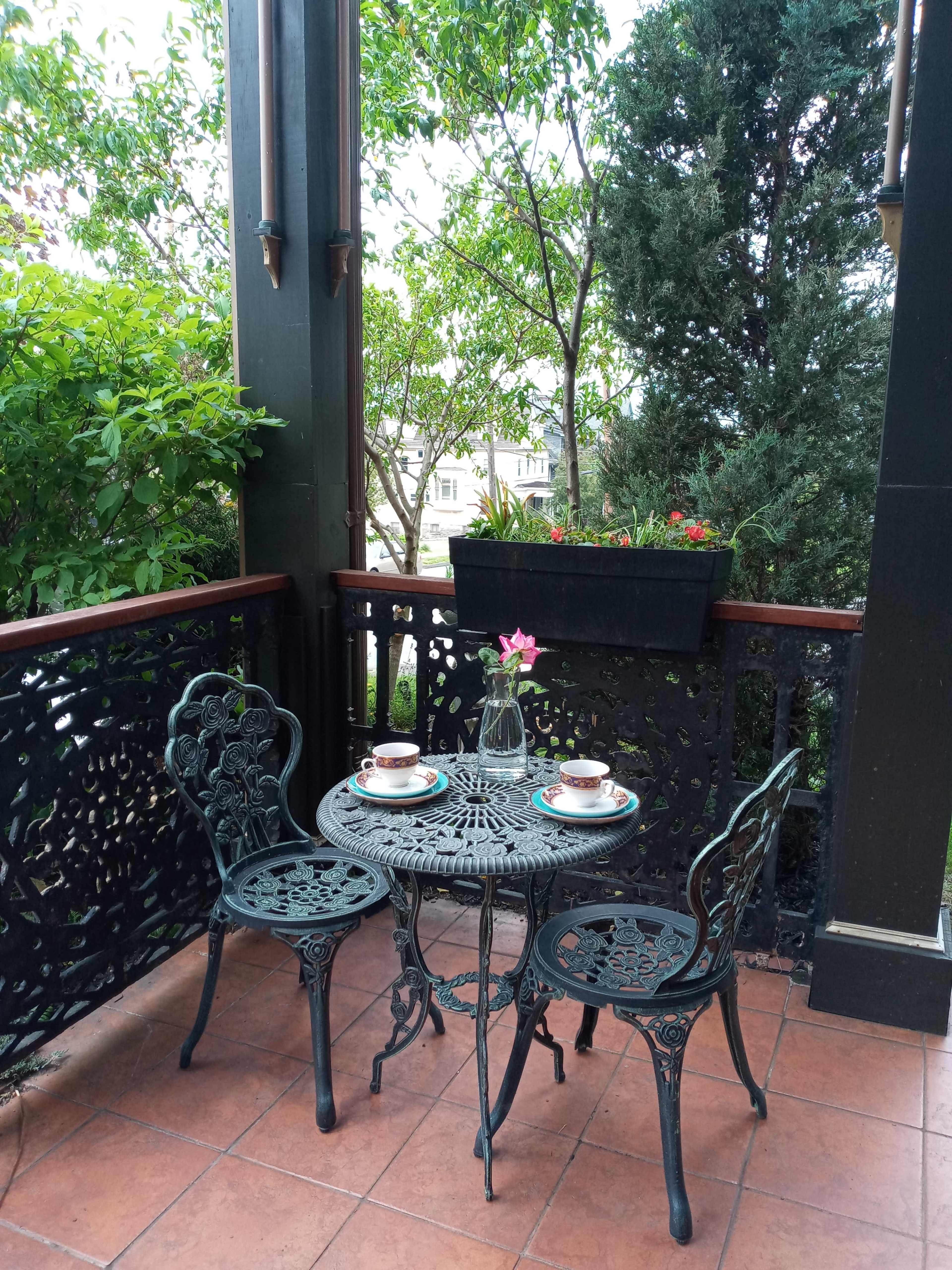 A small metal table with two chairs is set on a balcony surrounded by greenery, featuring two cups and a flower in the center.