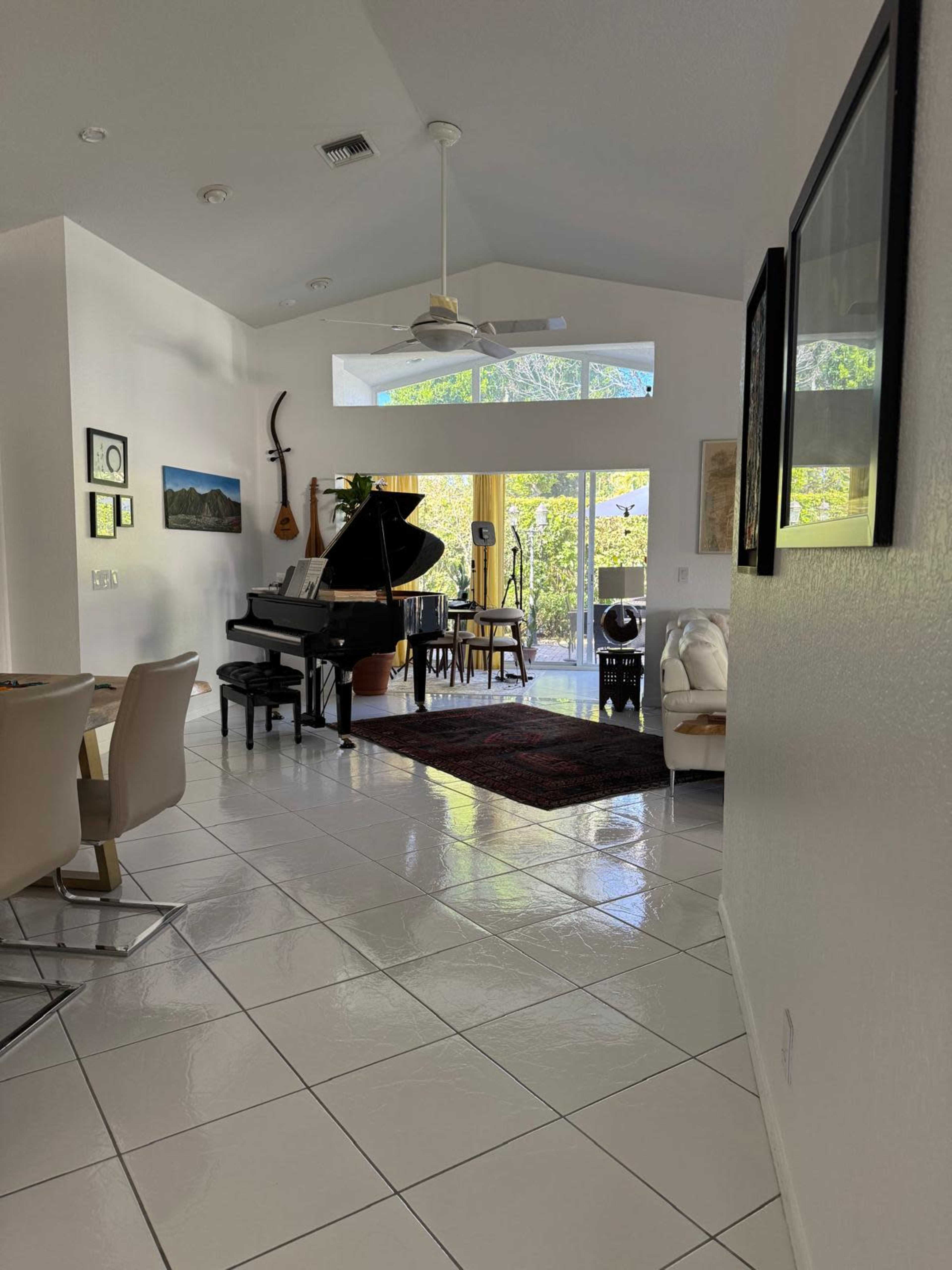 The image depicts a bright, open living space featuring a grand piano, dining area, and large windows overlooking greenery.