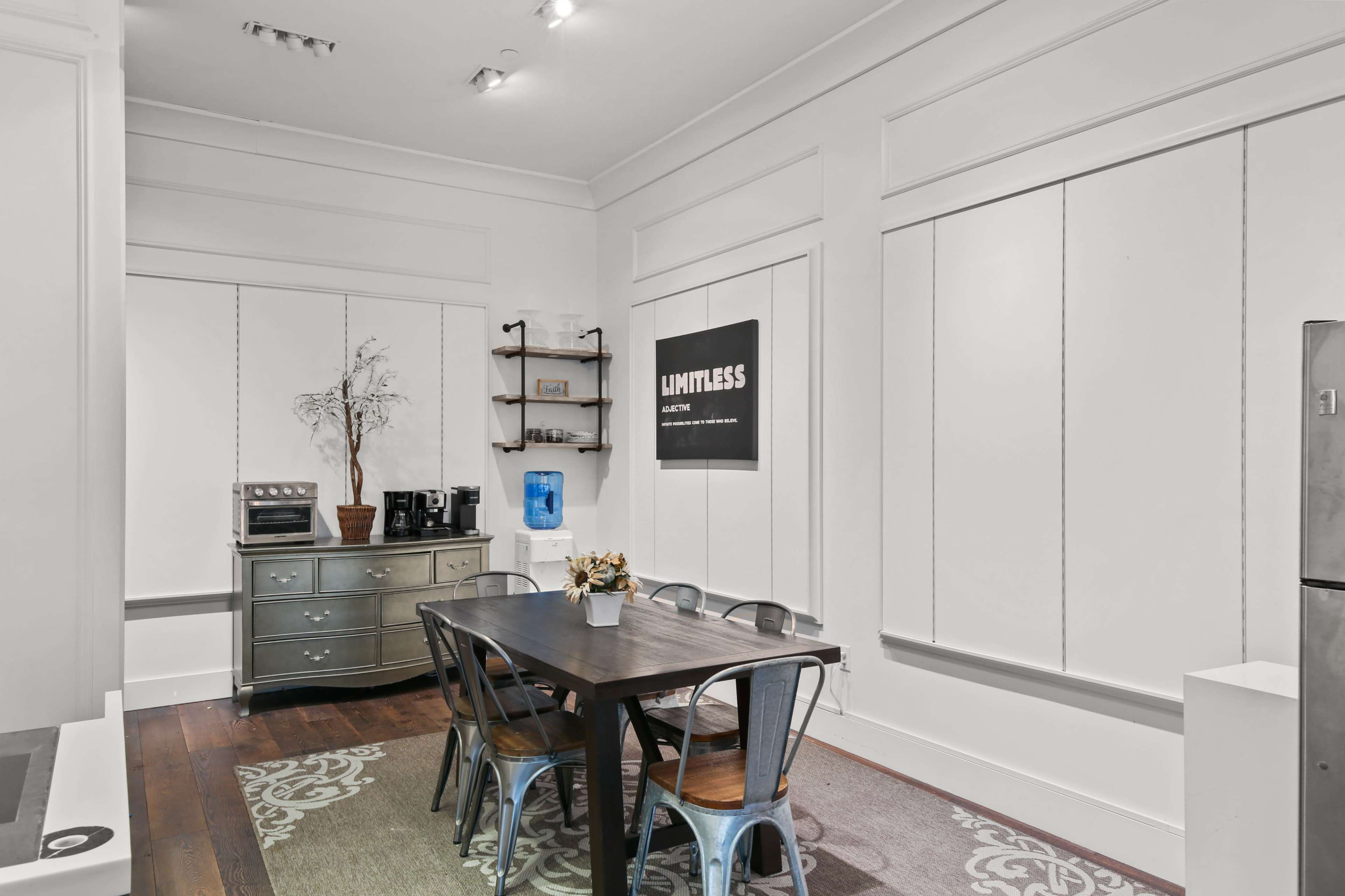 A dining area features a wooden table surrounded by metal chairs, with a water cooler, a silver dresser, and a large motivational poster on the wall.