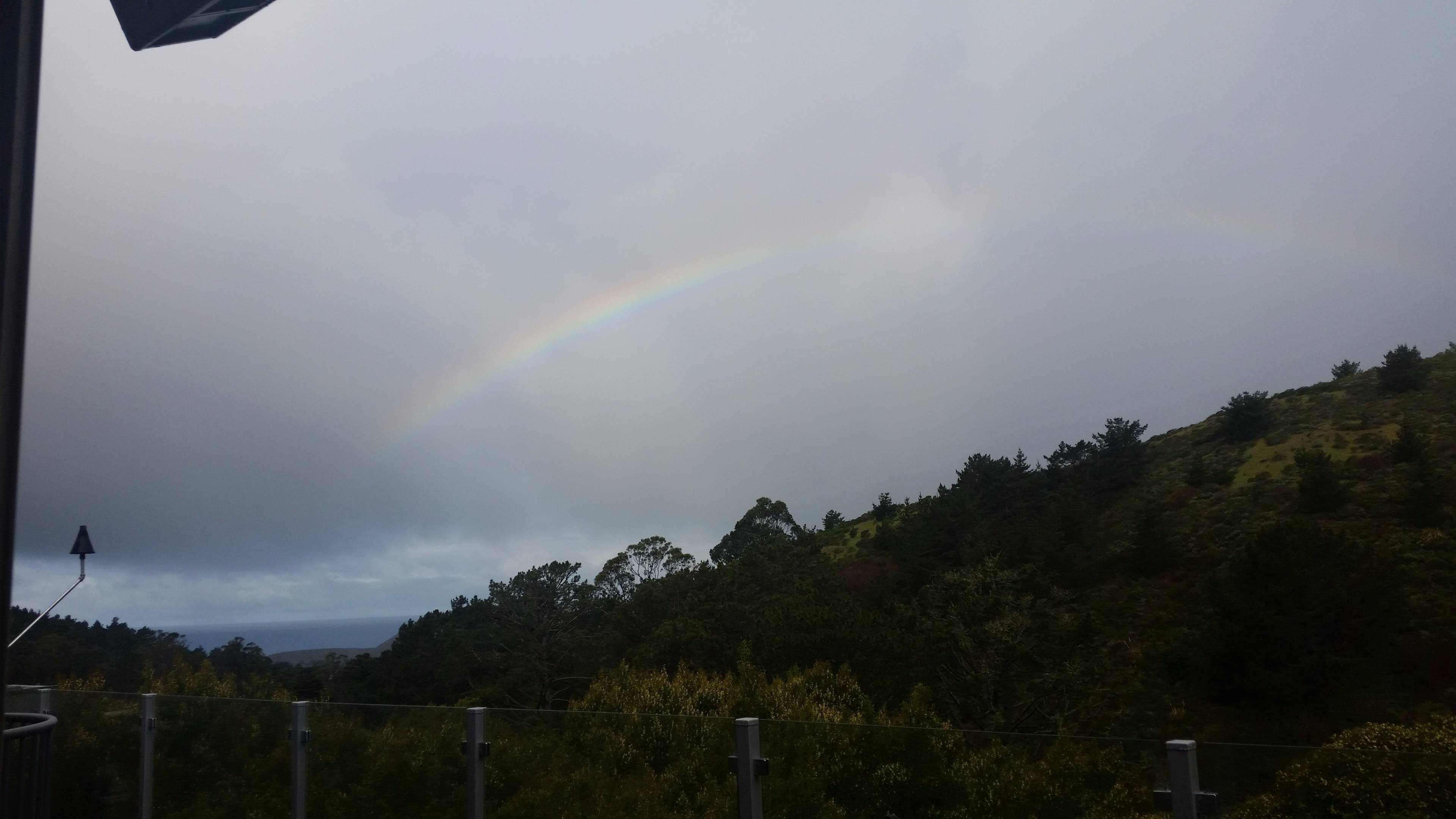 A rainbow arcs across a cloudy sky above a forested landscape.