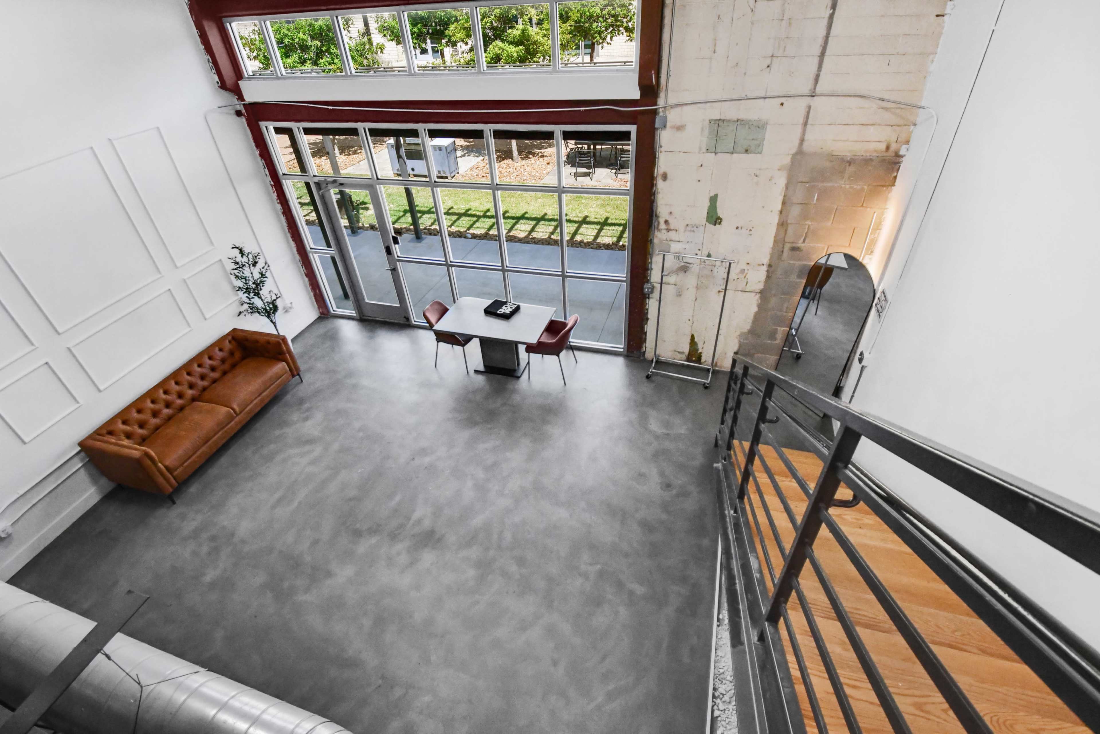 The image shows a spacious, modern room with a leather sofa, a table with chairs, large windows allowing natural light, and a staircase leading to a higher level.
