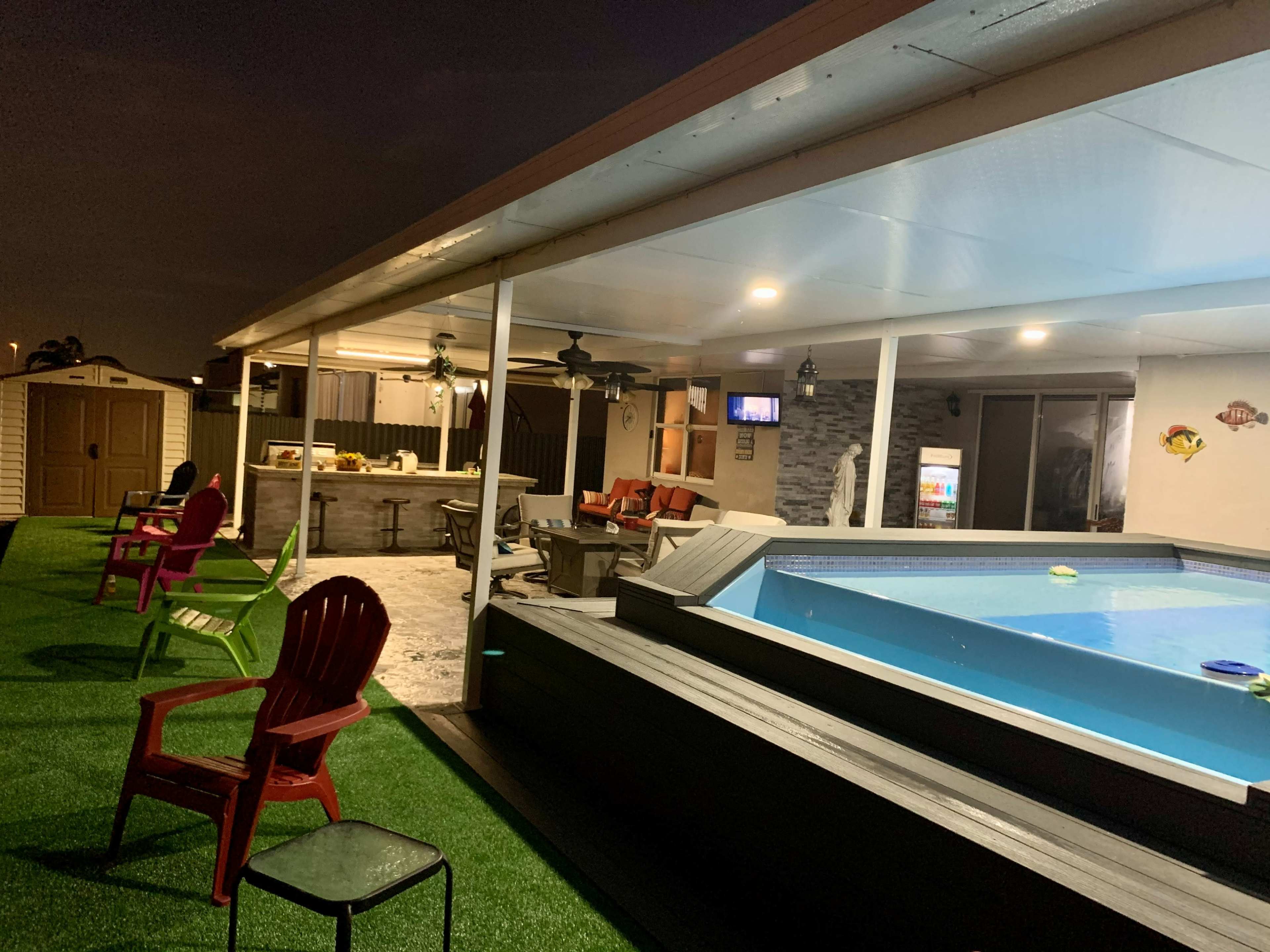 A backyard scene features a pool surrounded by seating areas, outdoor furniture, and a covered patio, illuminated by evening lights.