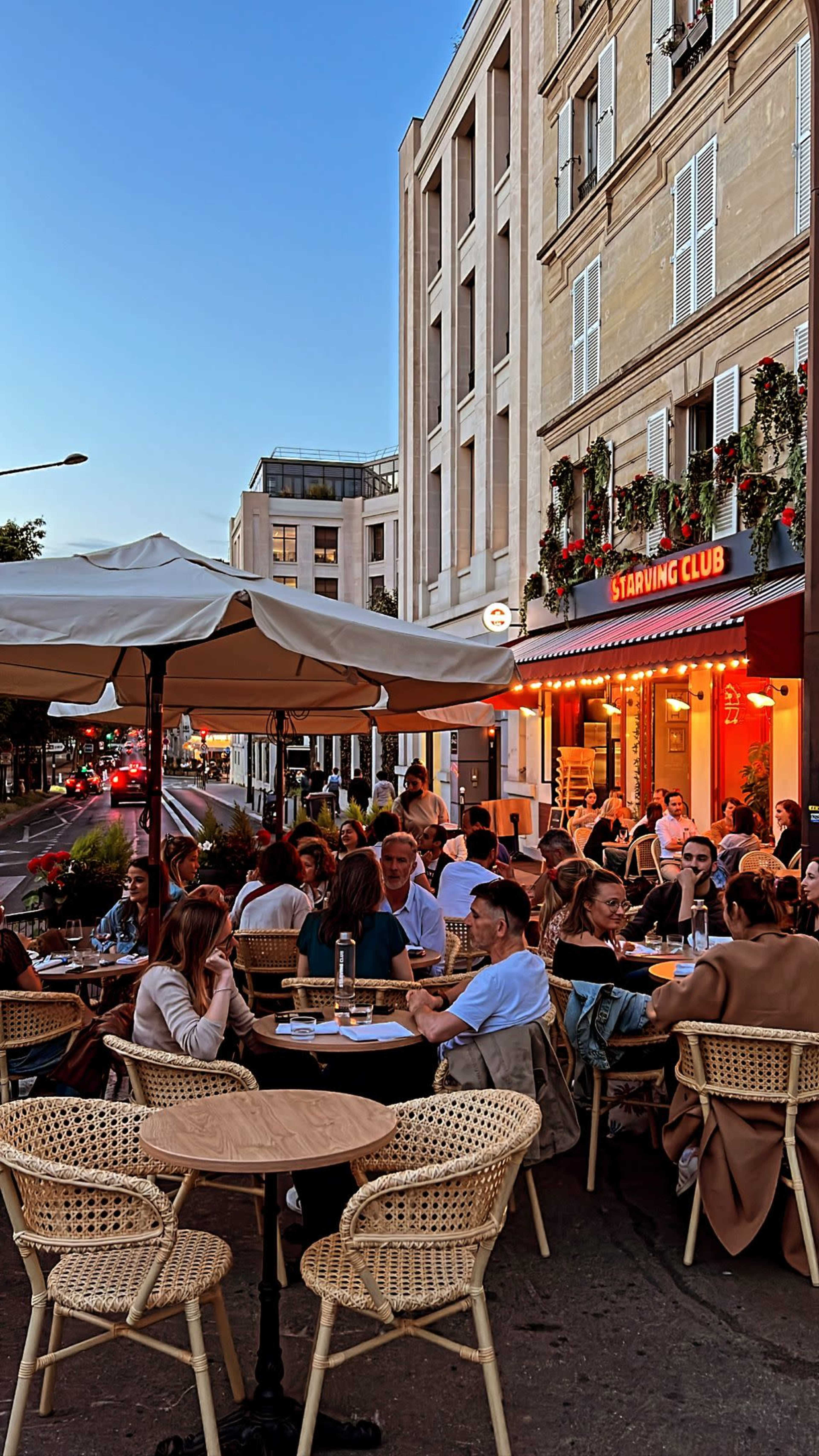 A busy street-side restaurant patio is filled with diners enjoying the evening atmosphere under string lights and nearby buildings.
