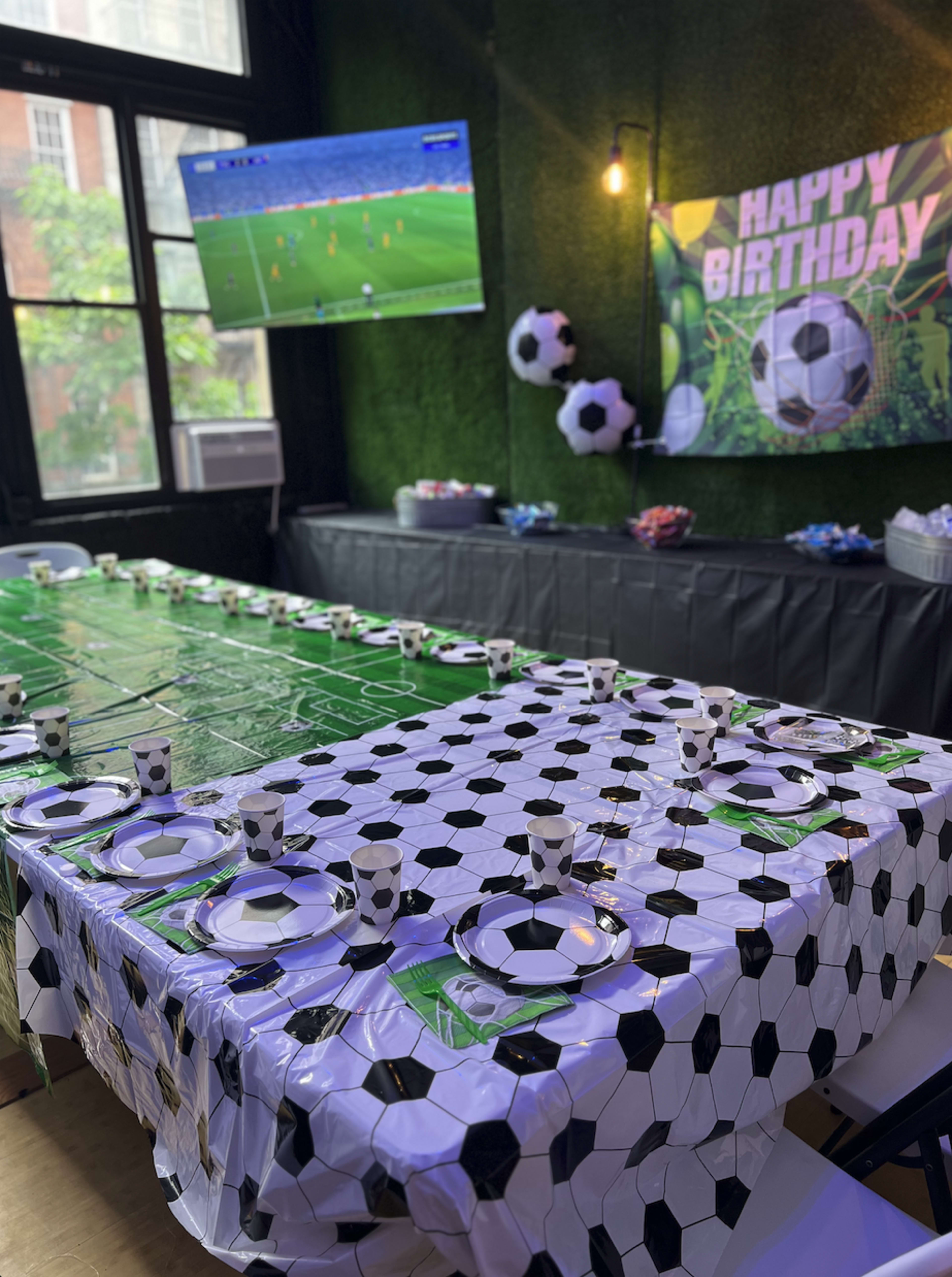 The scene features a birthday party setup with a green and black soccer-themed tablecloth, plastic cups, and plates, complemented by a large TV displaying a soccer game and decorative soccer balls on the walls.