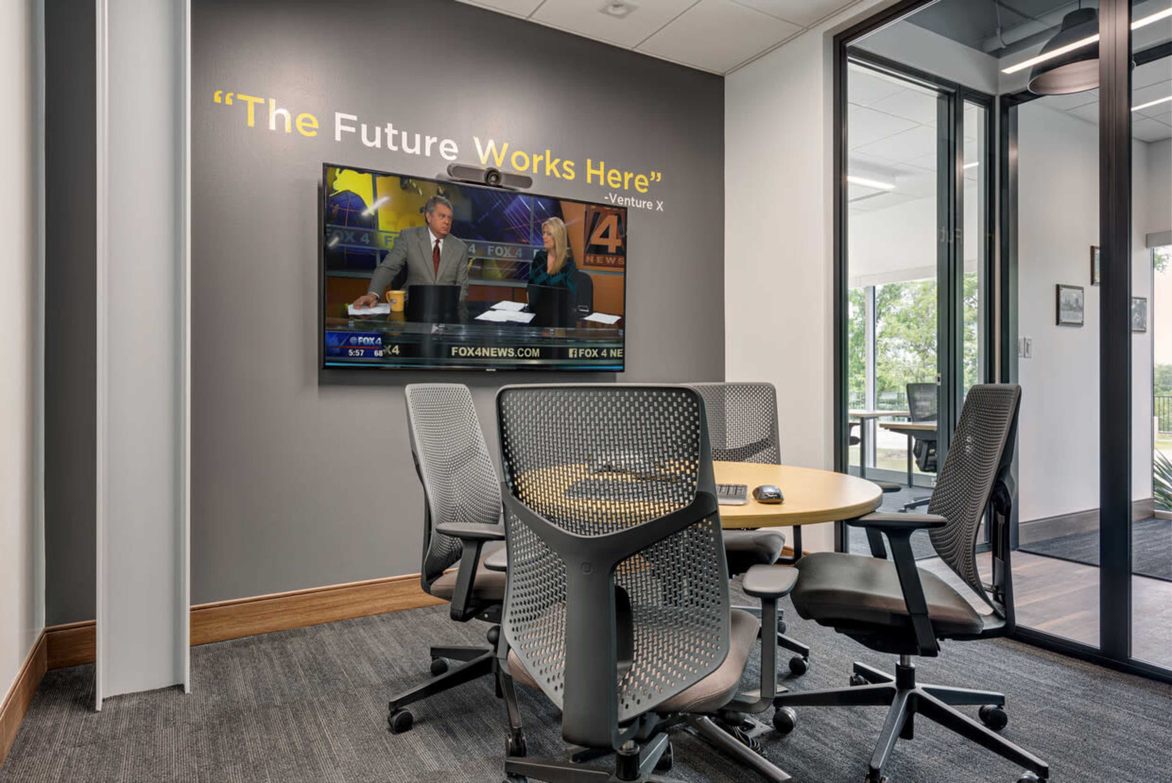 A conference room features a round table surrounded by ergonomic chairs, with a television displaying a news broadcast mounted on the wall.