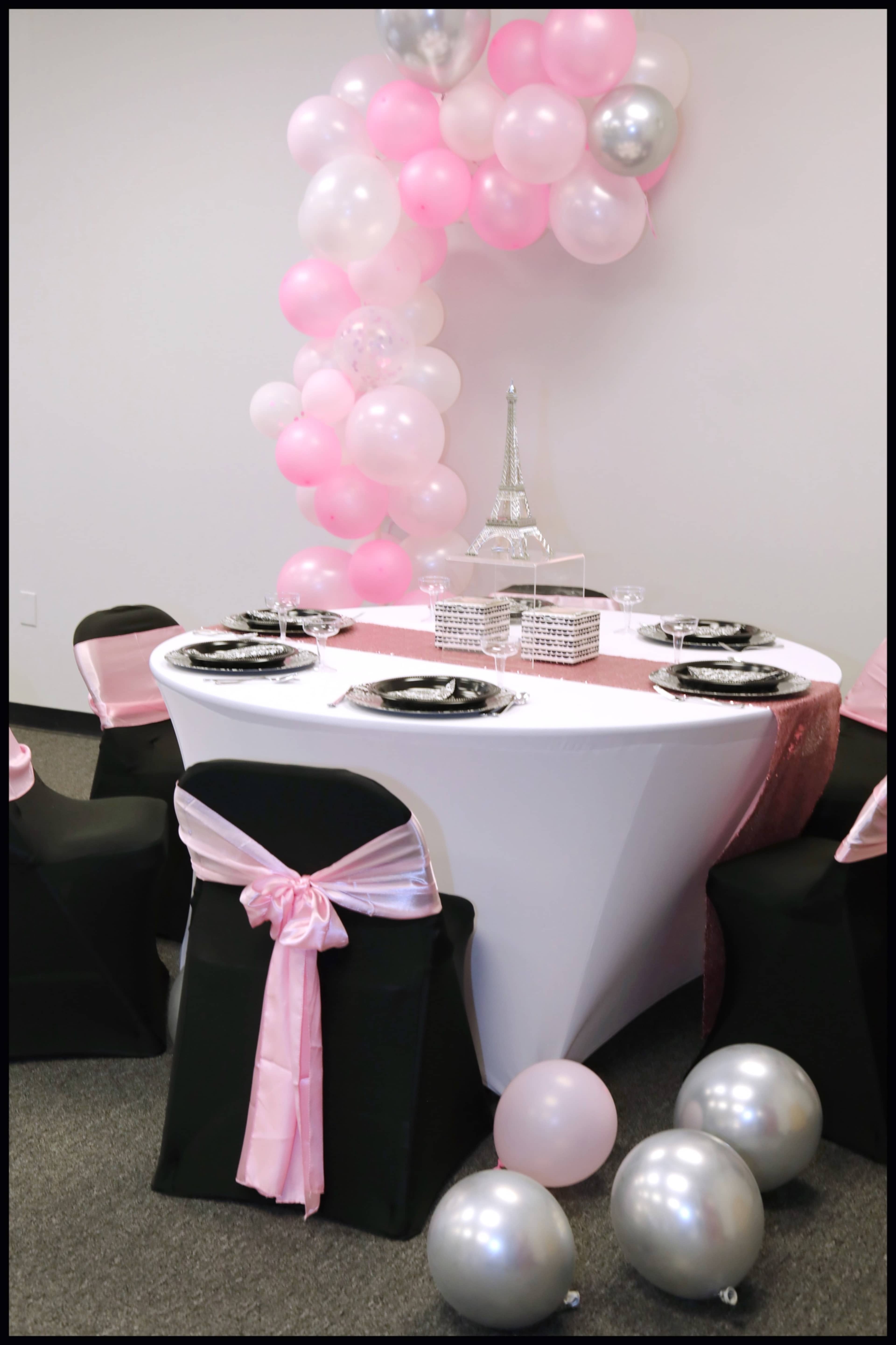 A round table with a pink and white table runner is set for a celebration, decorated with black chairs featuring pink sashes and surrounded by a cluster of balloons.