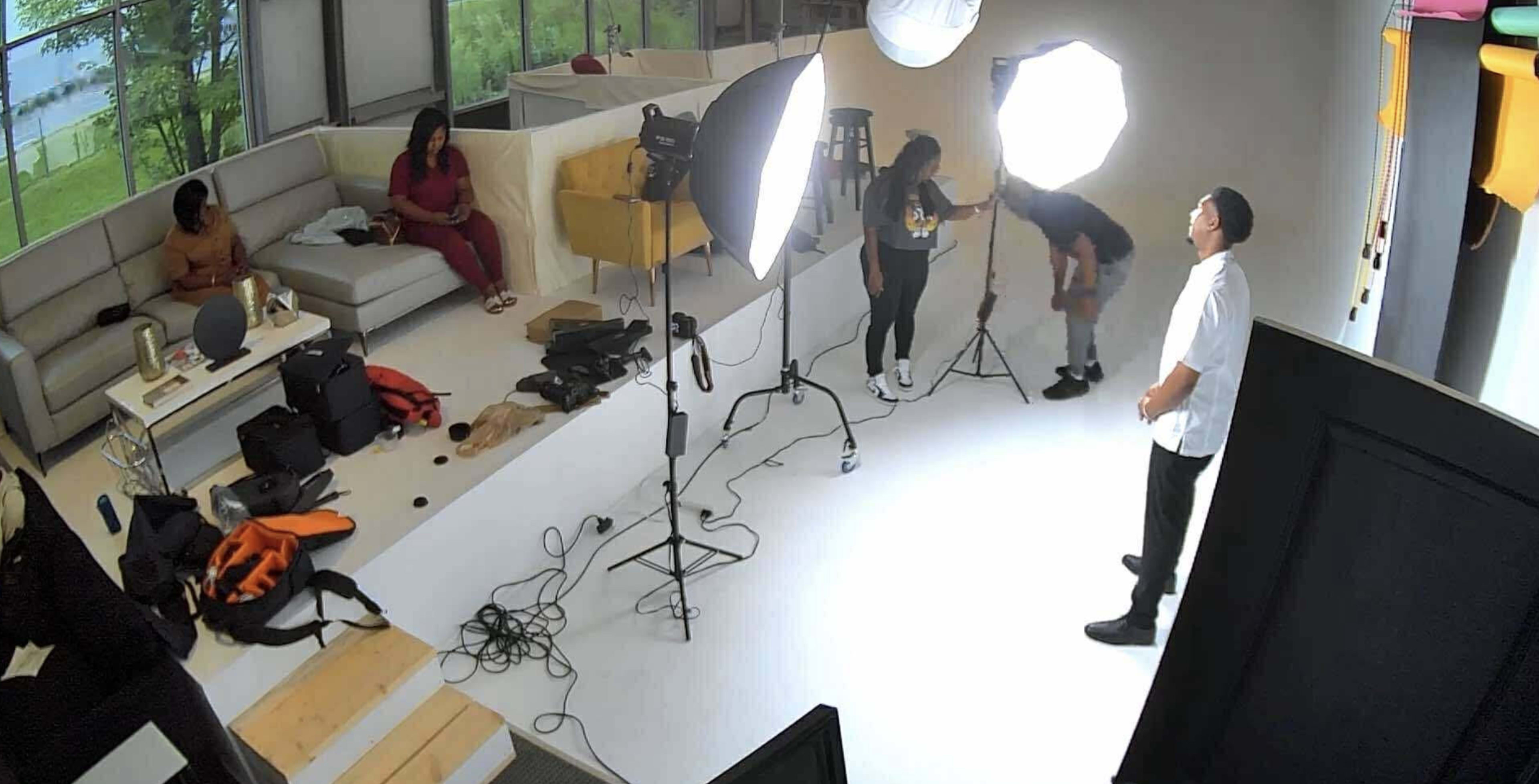 A photography studio is set up with bright lights, a subject posing in front of the camera, and a few people working in the background.