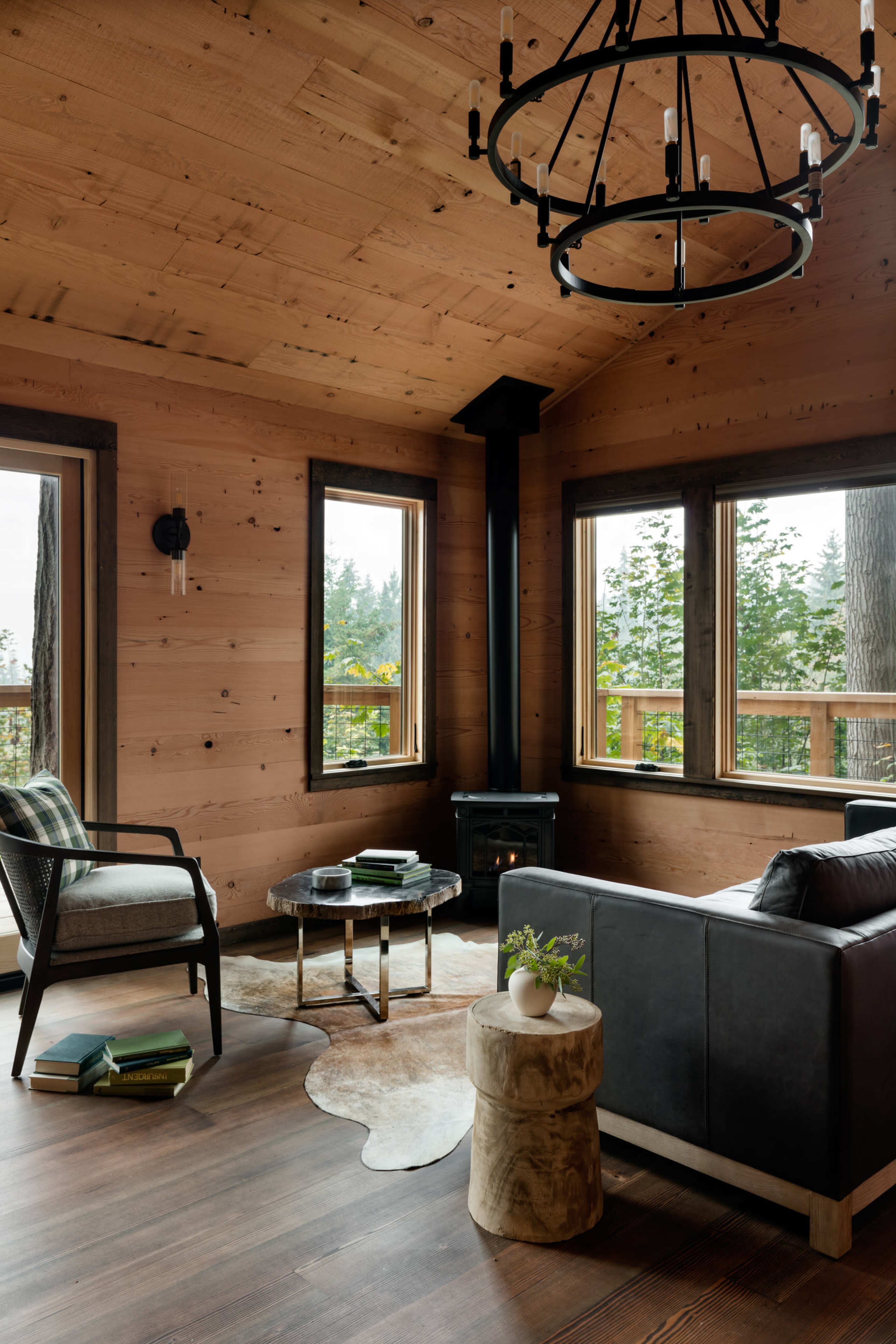 A cozy living room with wooden walls, a modern black sofa, a round coffee table, and large windows overlooking greenery.