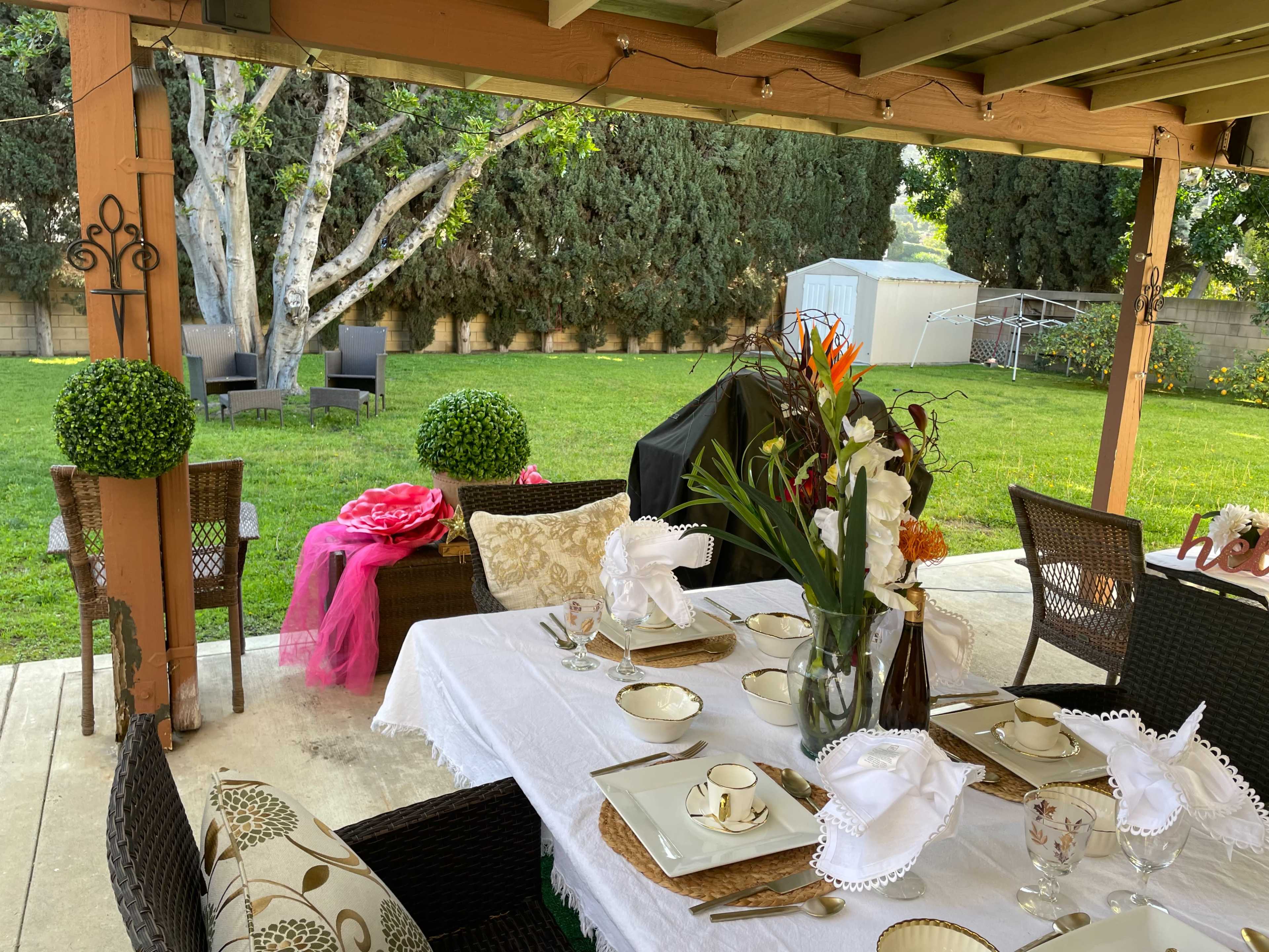 A well-set dining table is arranged under a covered patio, overlooking a green yard with garden chairs and neatly trimmed hedges.