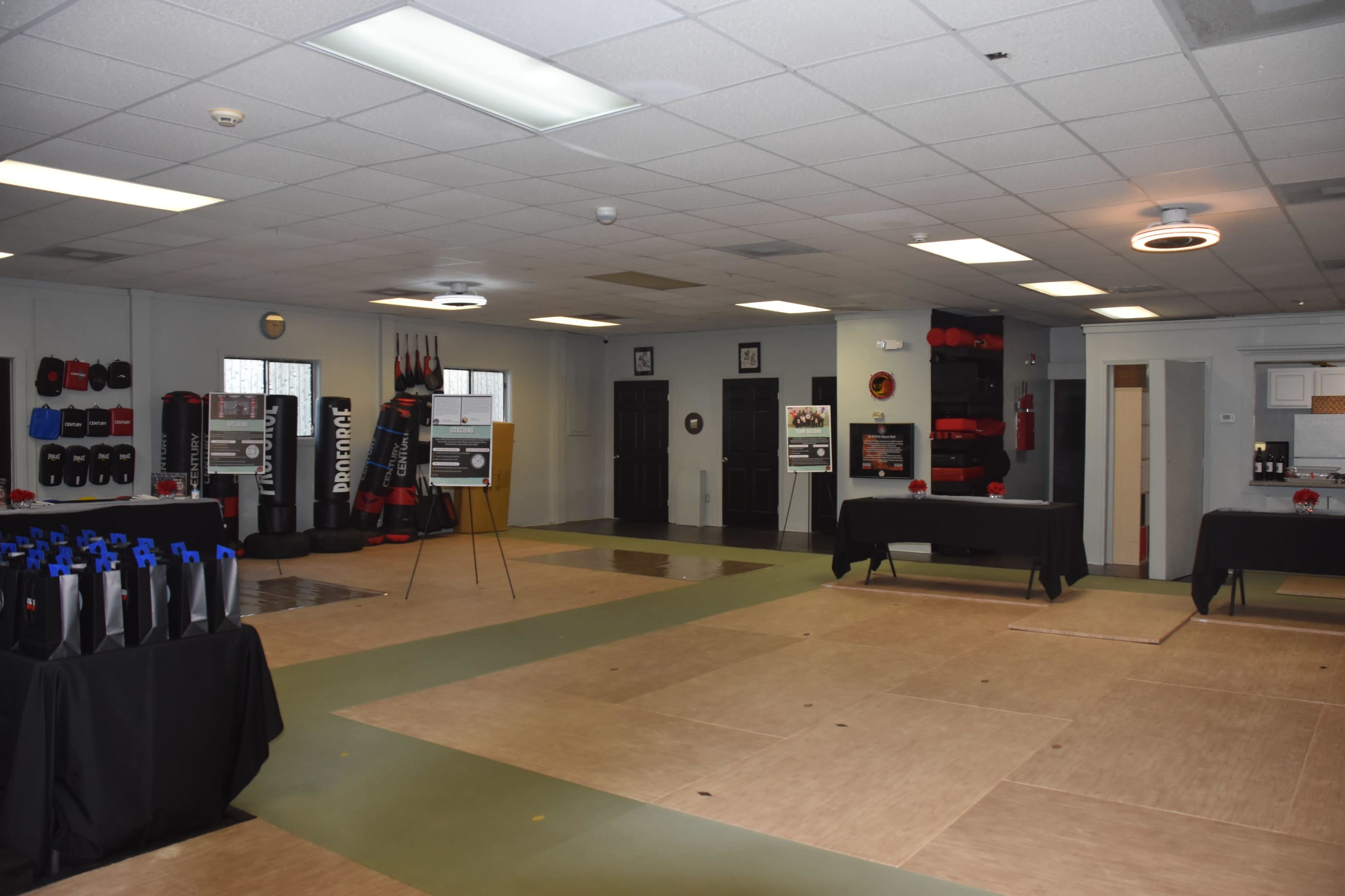 A spacious training room with wooden flooring, black tables, and various fitness equipment and boxing bags displayed along the walls.