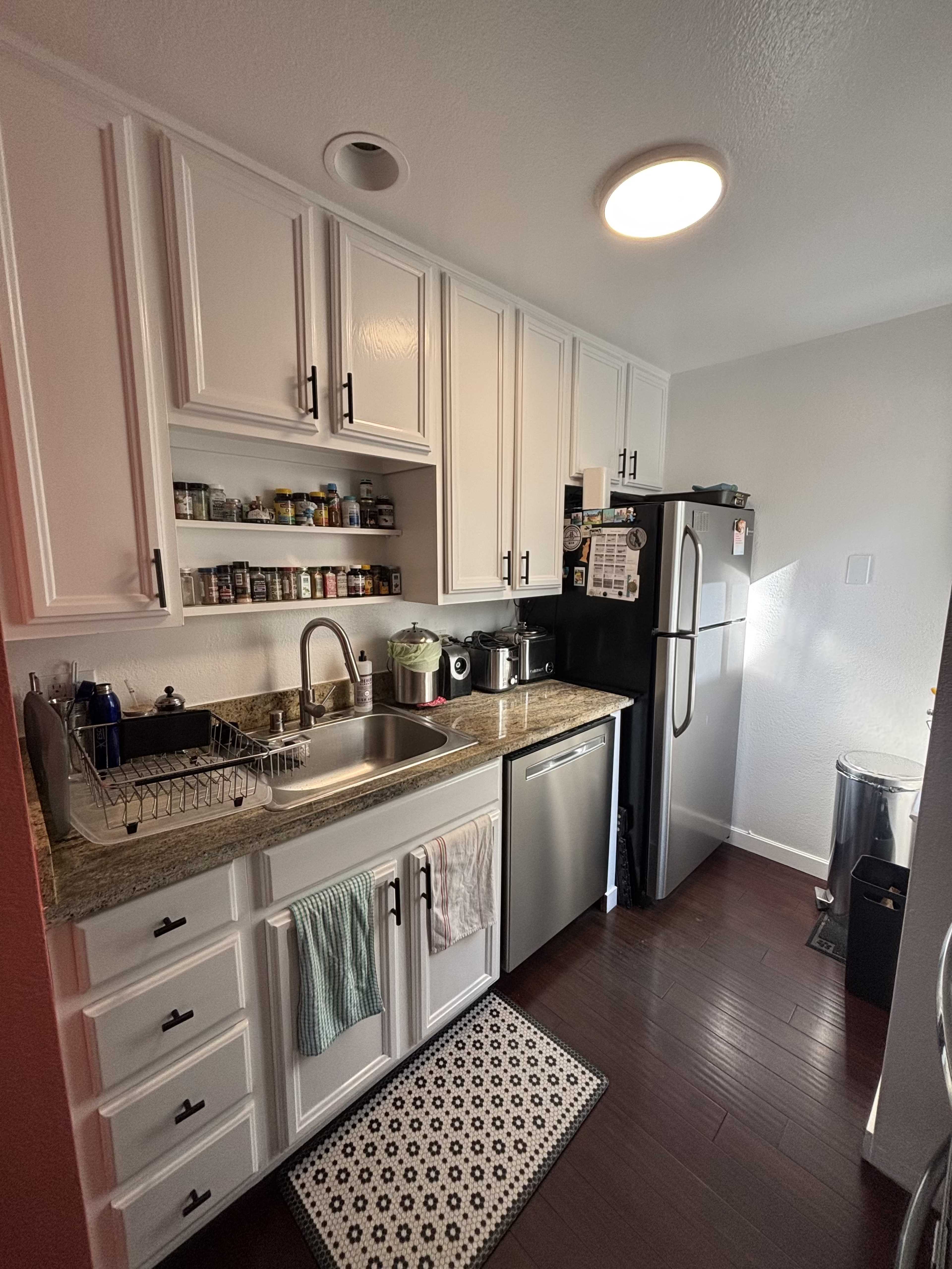 A compact kitchen features white cabinetry, a stainless steel sink, a black refrigerator, and various kitchen supplies organized on shelves and countertops.