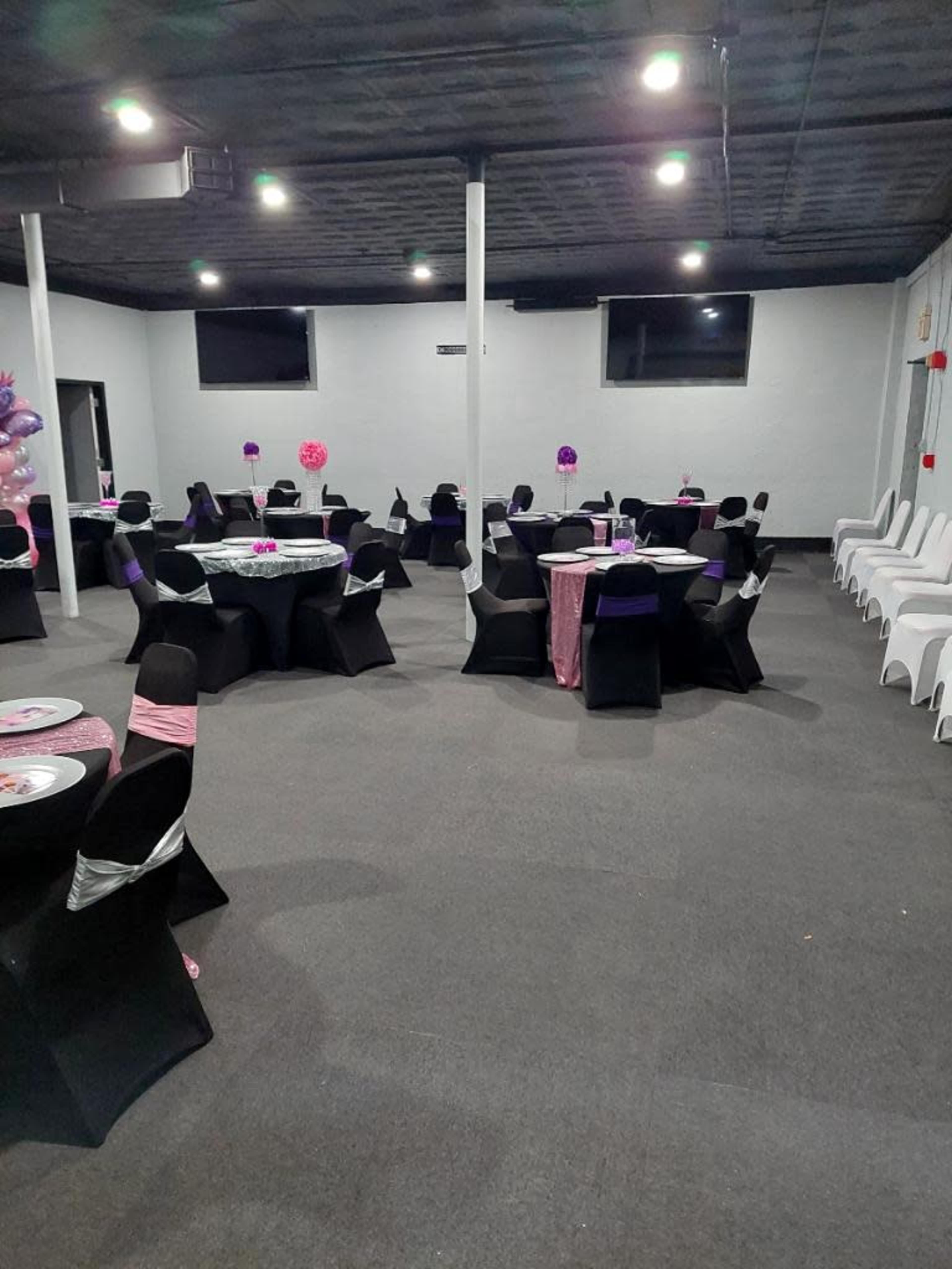 The image shows a party venue with several round tables set with black and pink decorations, surrounded by black and white chairs.