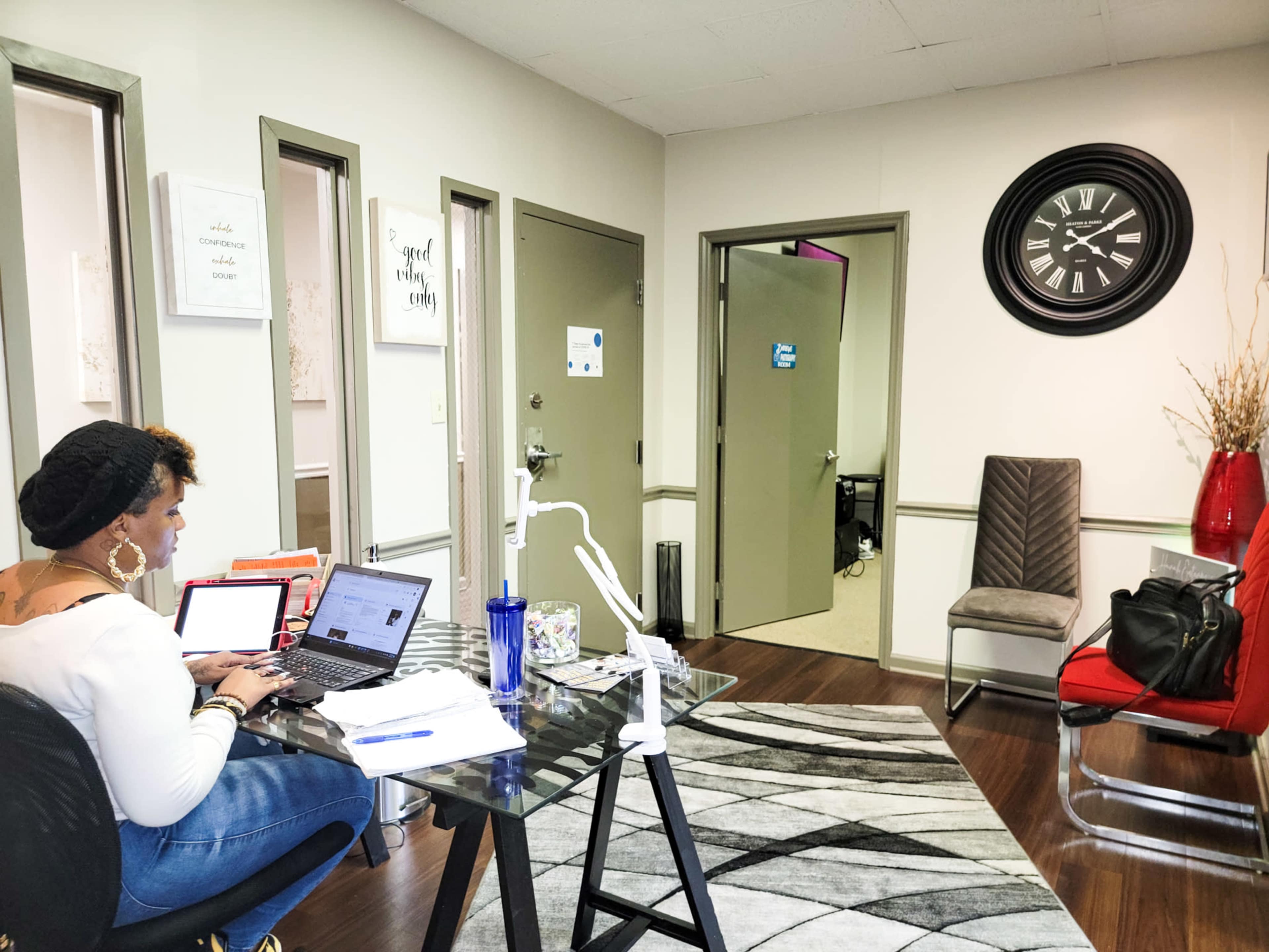 A woman works at a desk with a laptop in a modern office space featuring gray walls and a wall clock.