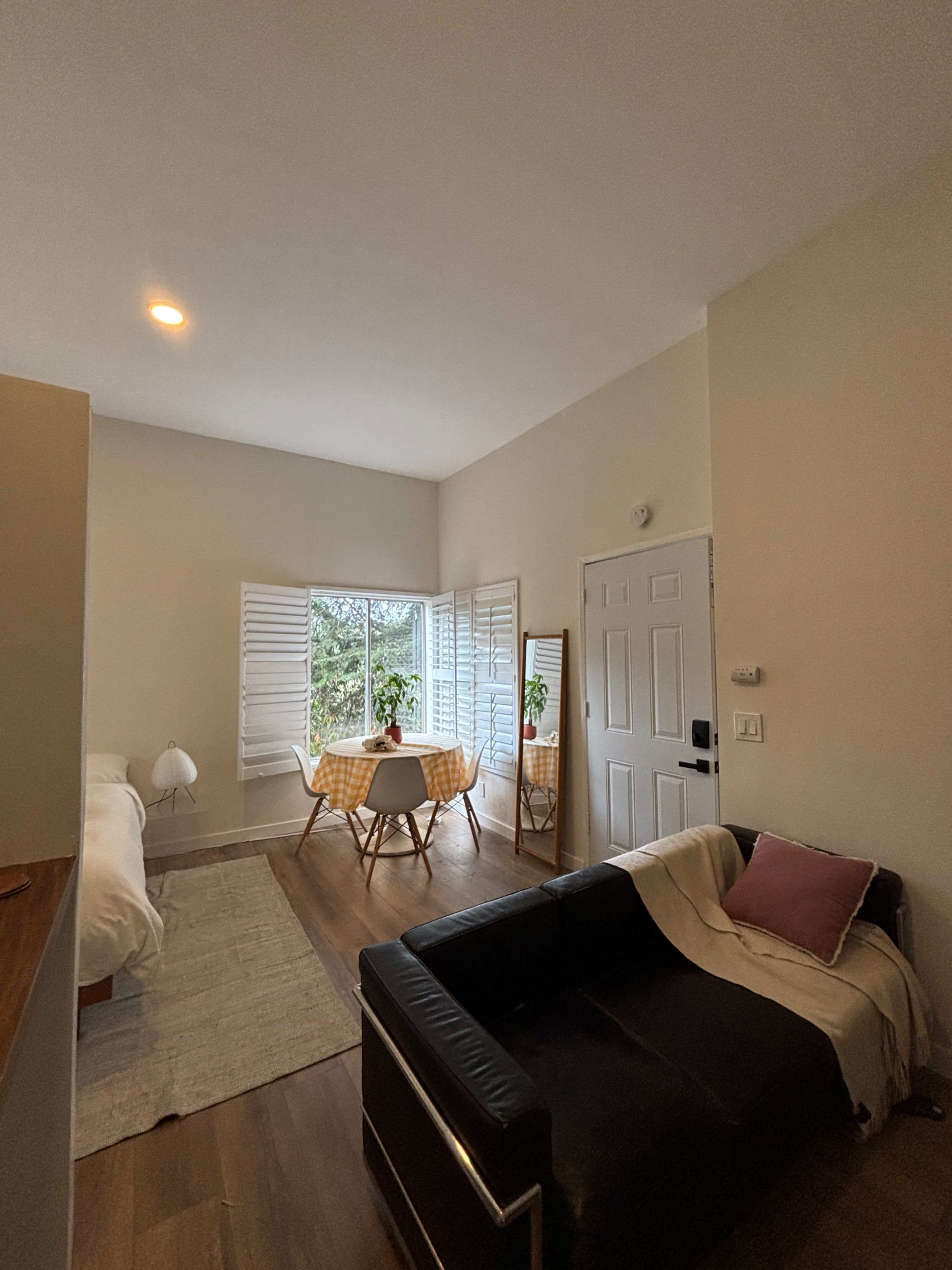 The image shows a small, bright room featuring a bed, a couch, and a dining table with chairs positioned near windows with shutters.