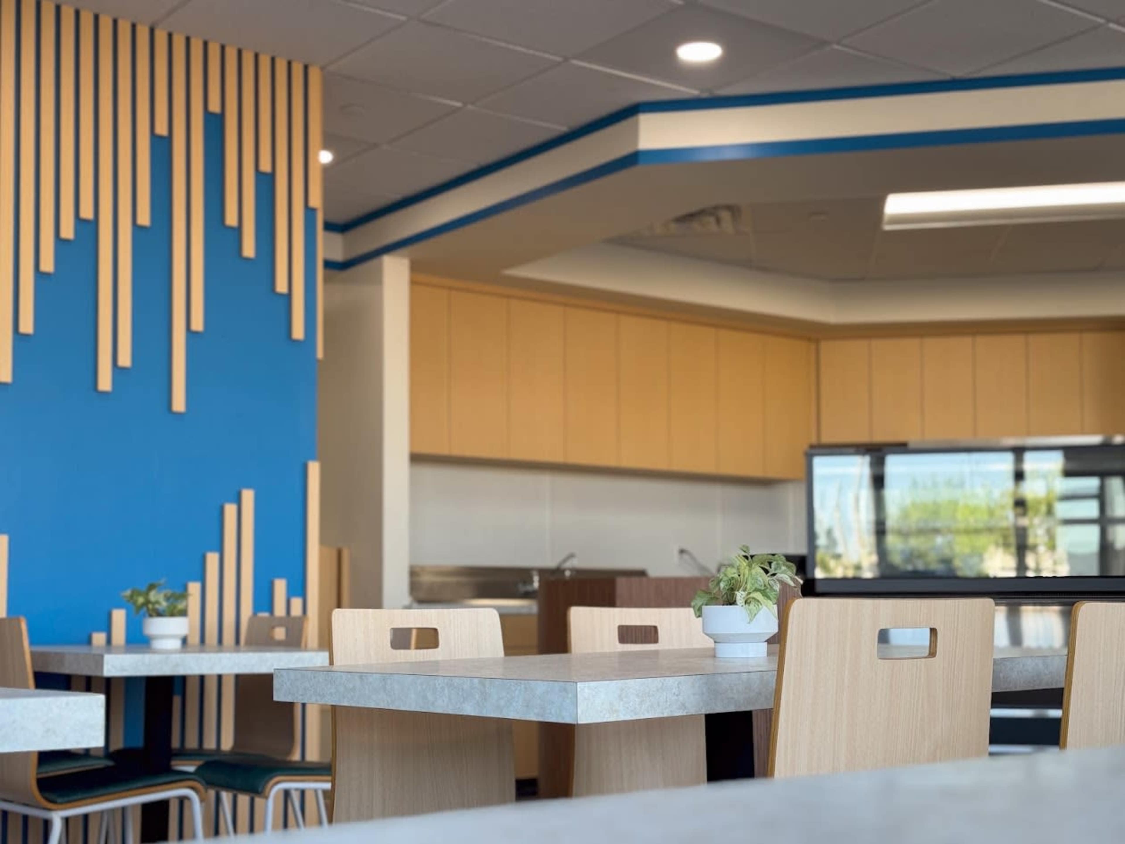 The image shows a modern dining area with light wood furniture and blue and wooden wall decor behind a kitchen space.