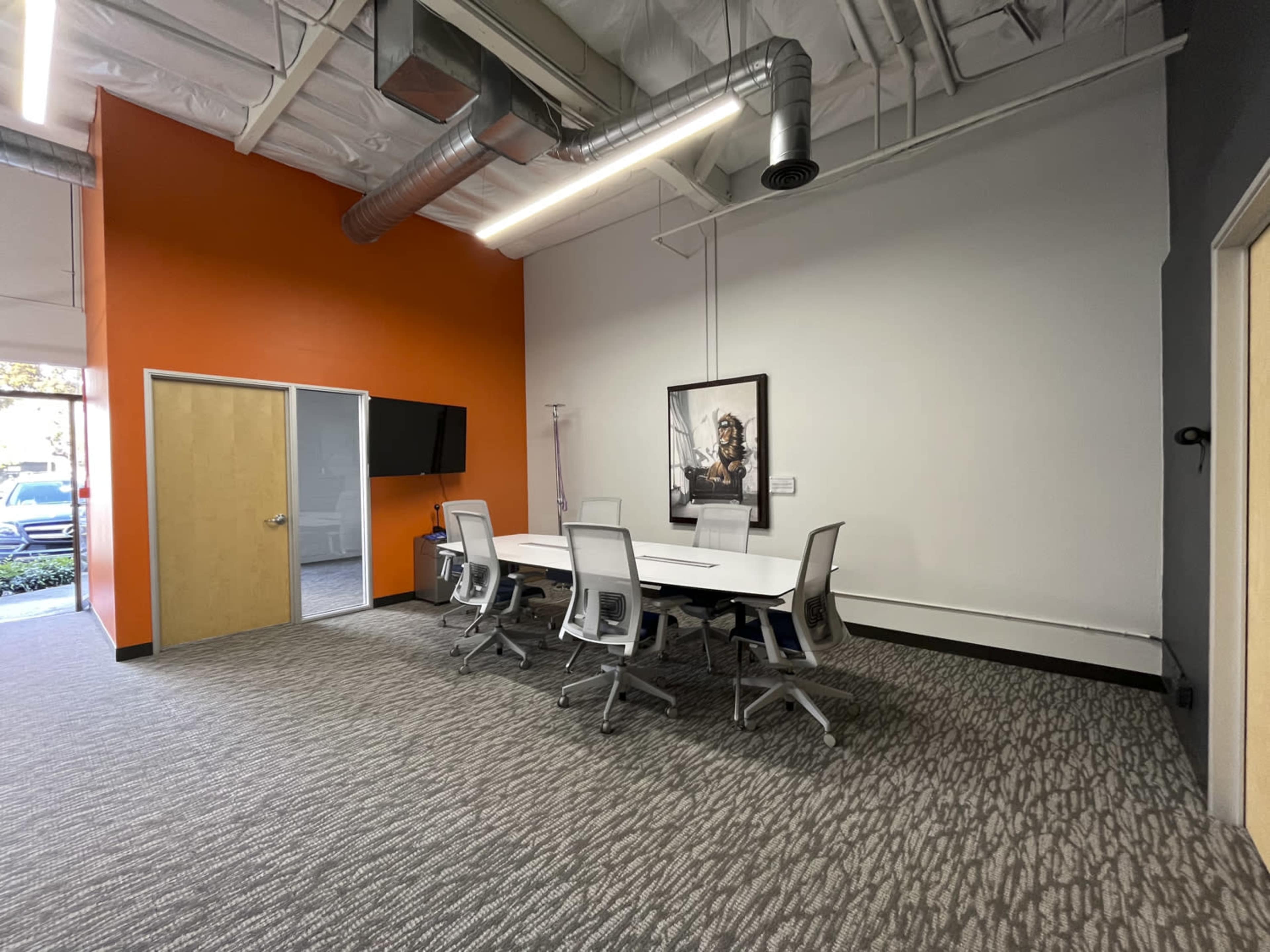 A conference room features a long white table surrounded by ergonomic chairs, with an orange accent wall and a framed artwork.