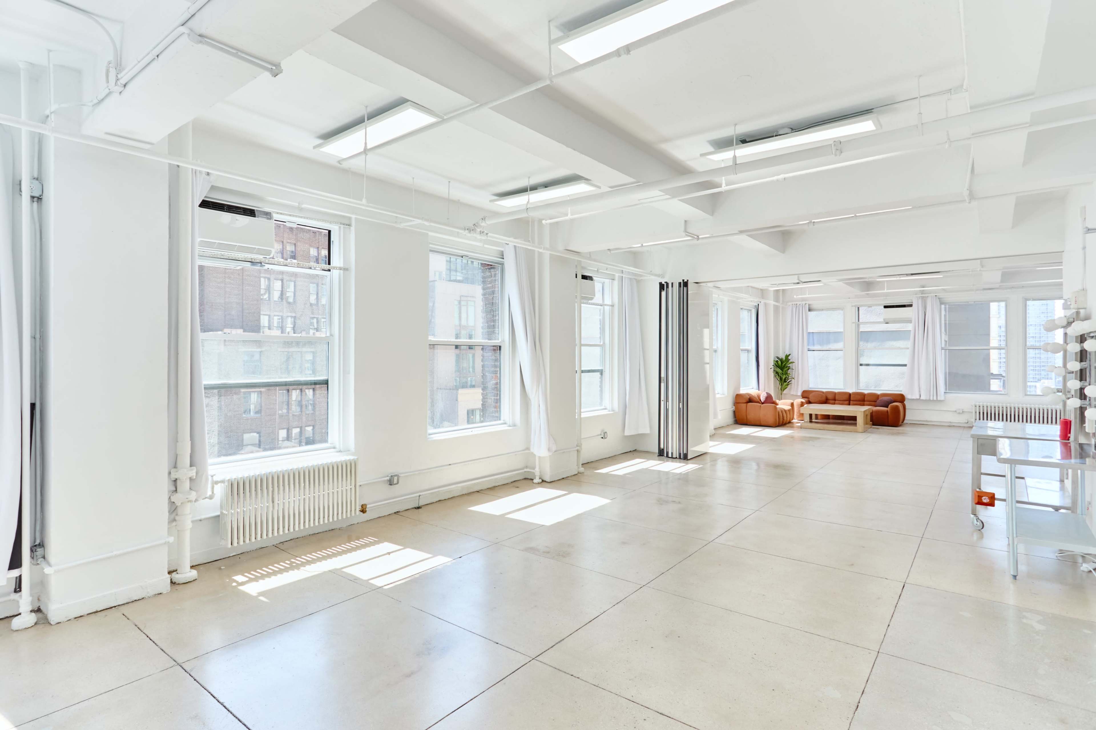 The image shows a spacious, bright room with large windows, white walls, and minimal furniture, including a leather couch and a small plant.