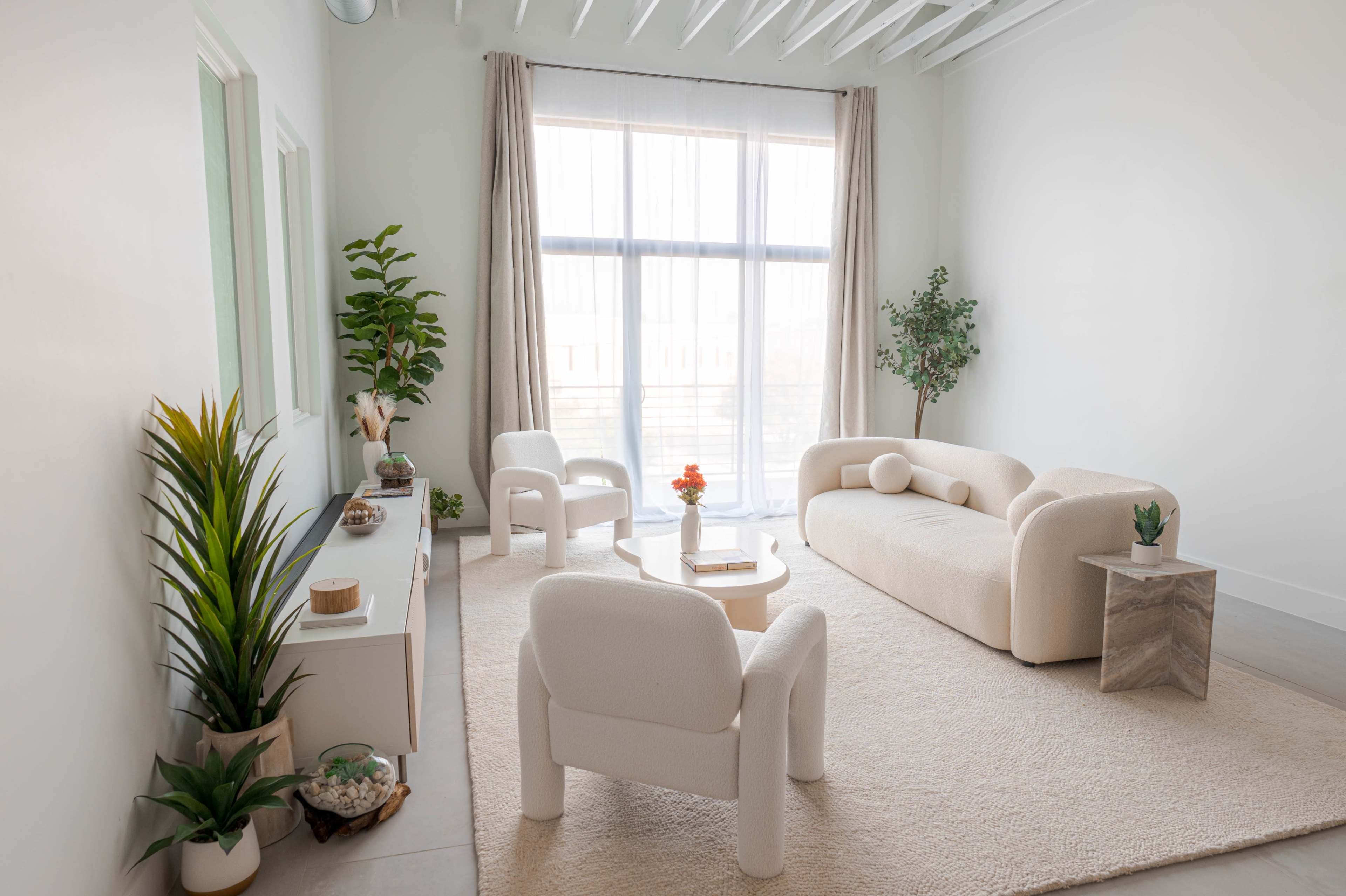 A light-filled living room features a minimalist design with a curved sofa, two armchairs, and a coffee table on a soft rug, complemented by green plants and neutral decor.