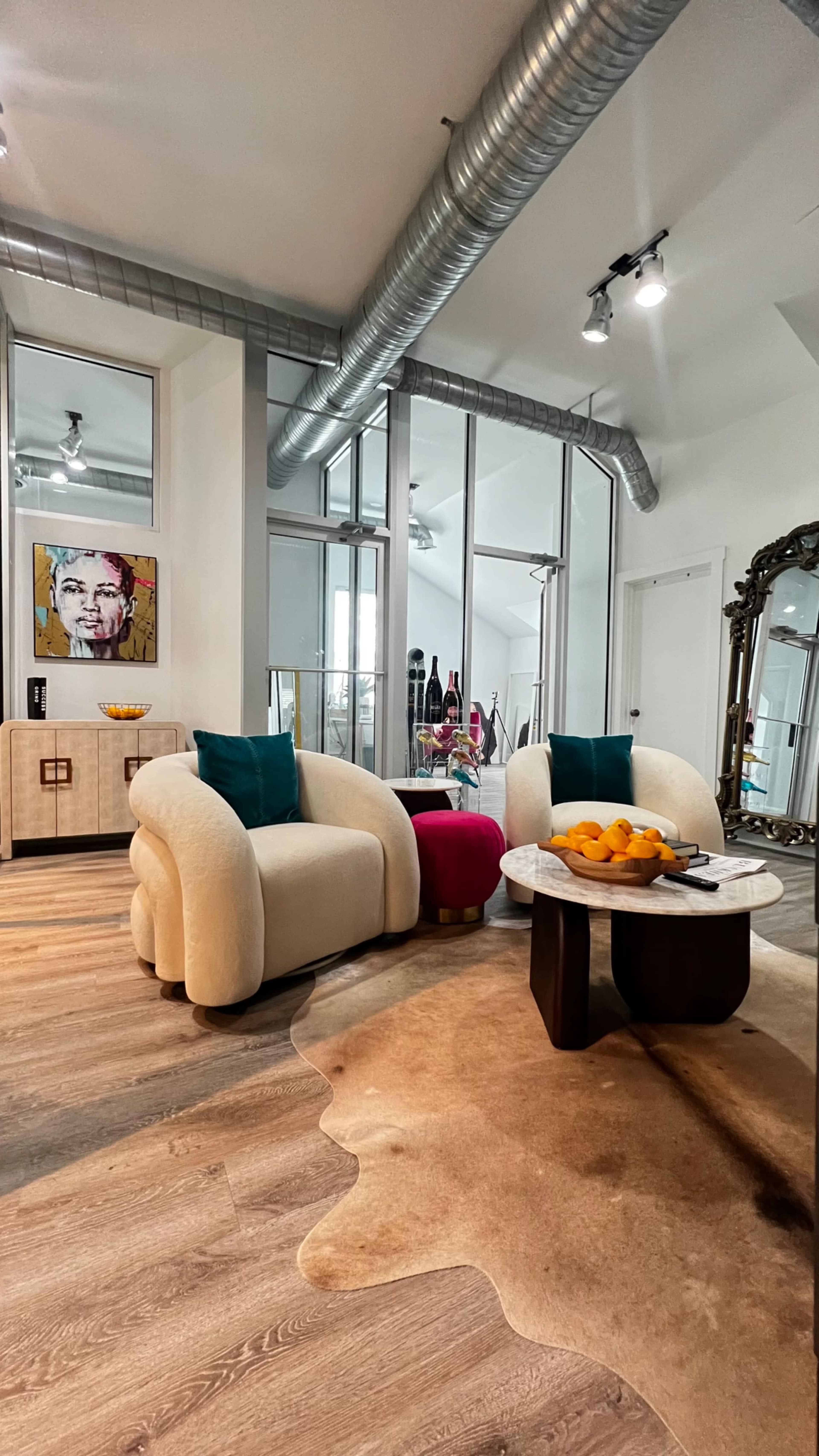 The image shows a modern living room featuring two curved, cream-colored chairs with teal cushions, a circular coffee table topped with oranges, and large windows that allow natural light to illuminate the space.